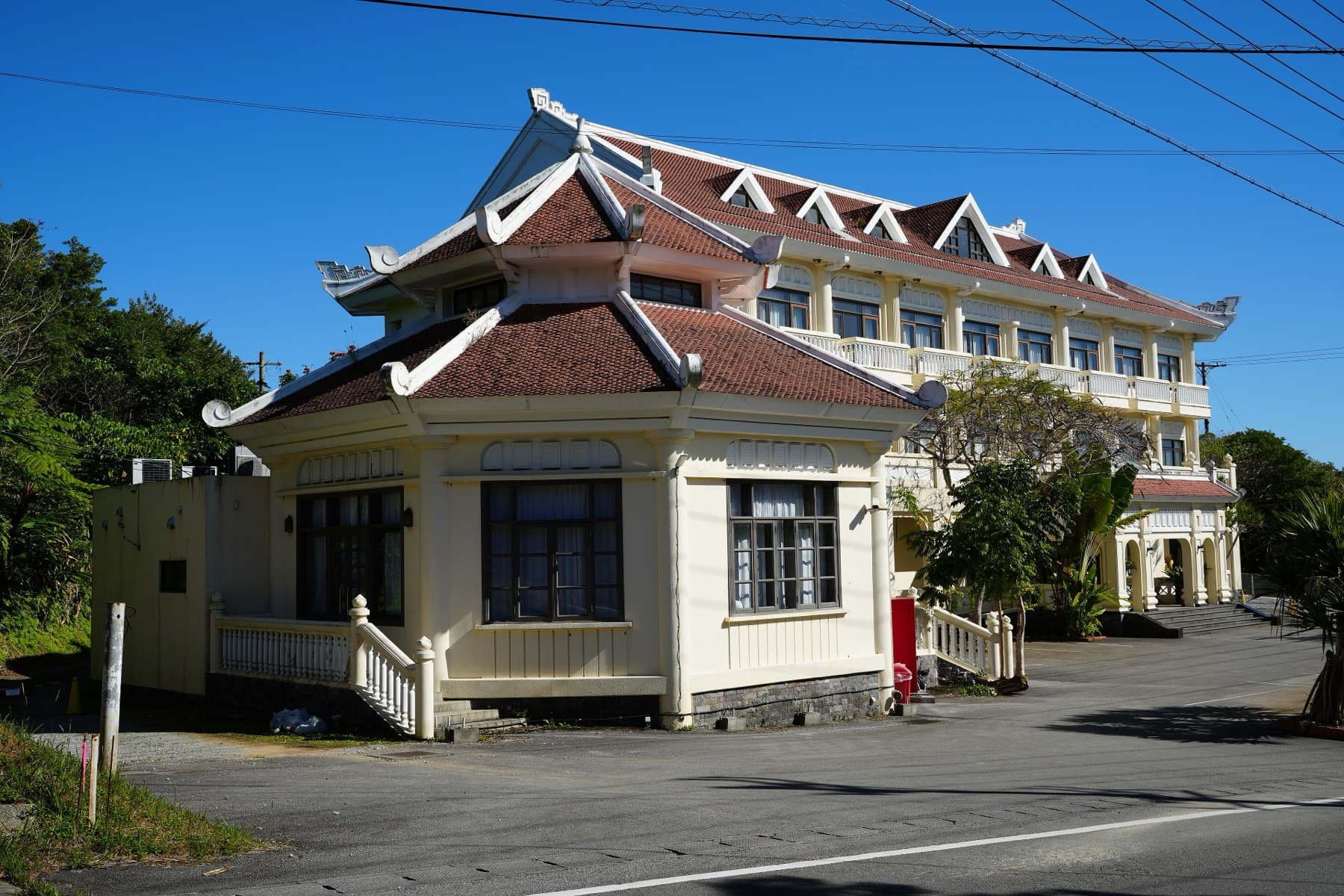 A cream hotel building with red tiled roofs and upturned eaves stands beside a quiet road in Kunigami.