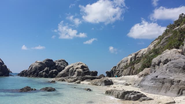 Rounded grey boulders crowd a small cove near Aharen on Tokashiki, and calm clear water fills the gaps between them under a high blue sky