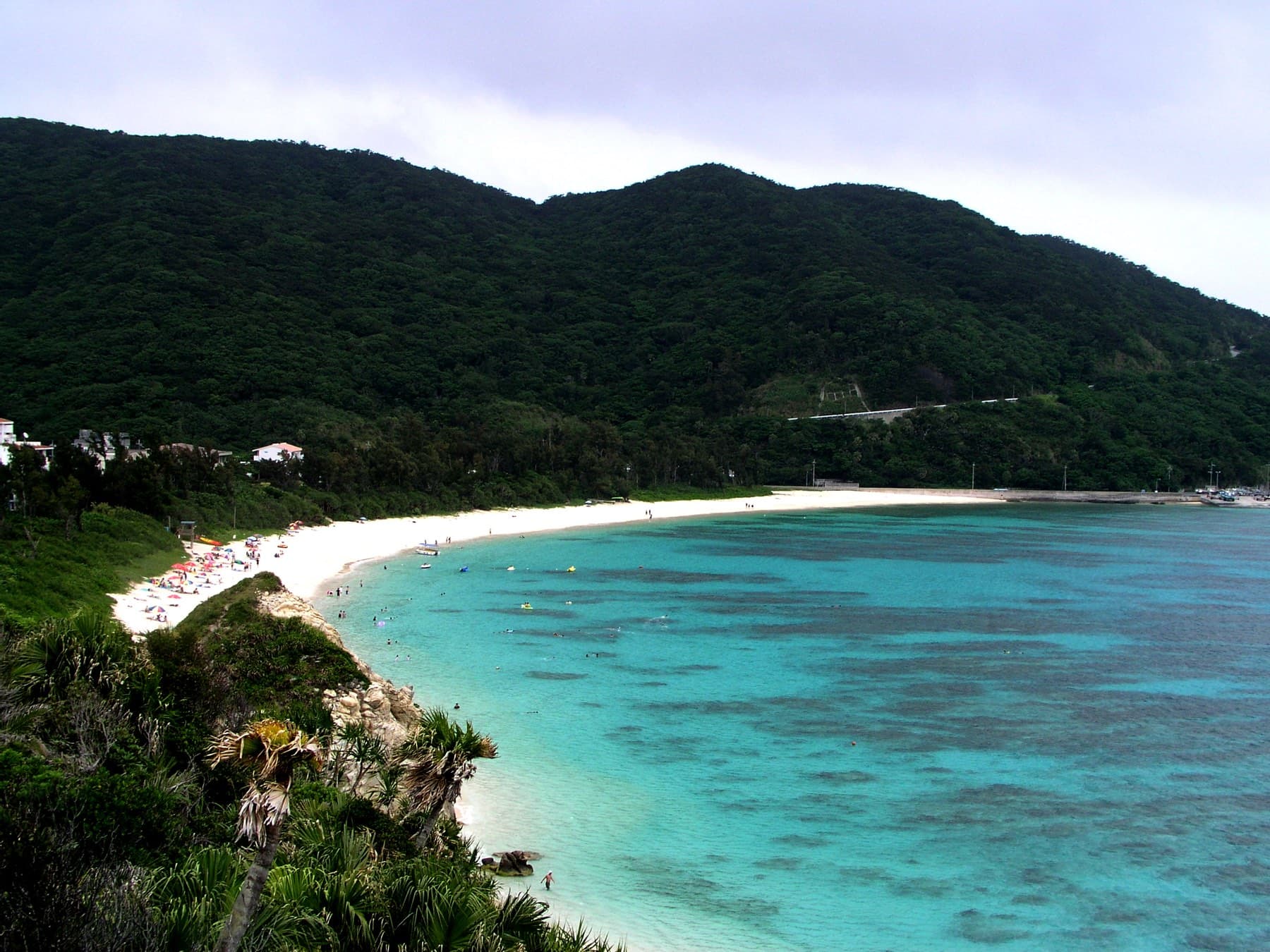 Under a grey sky the shallows at Aharen Beach on Tokashiki still glow pale blue over the reef, and parasols dot the sand below forested hills