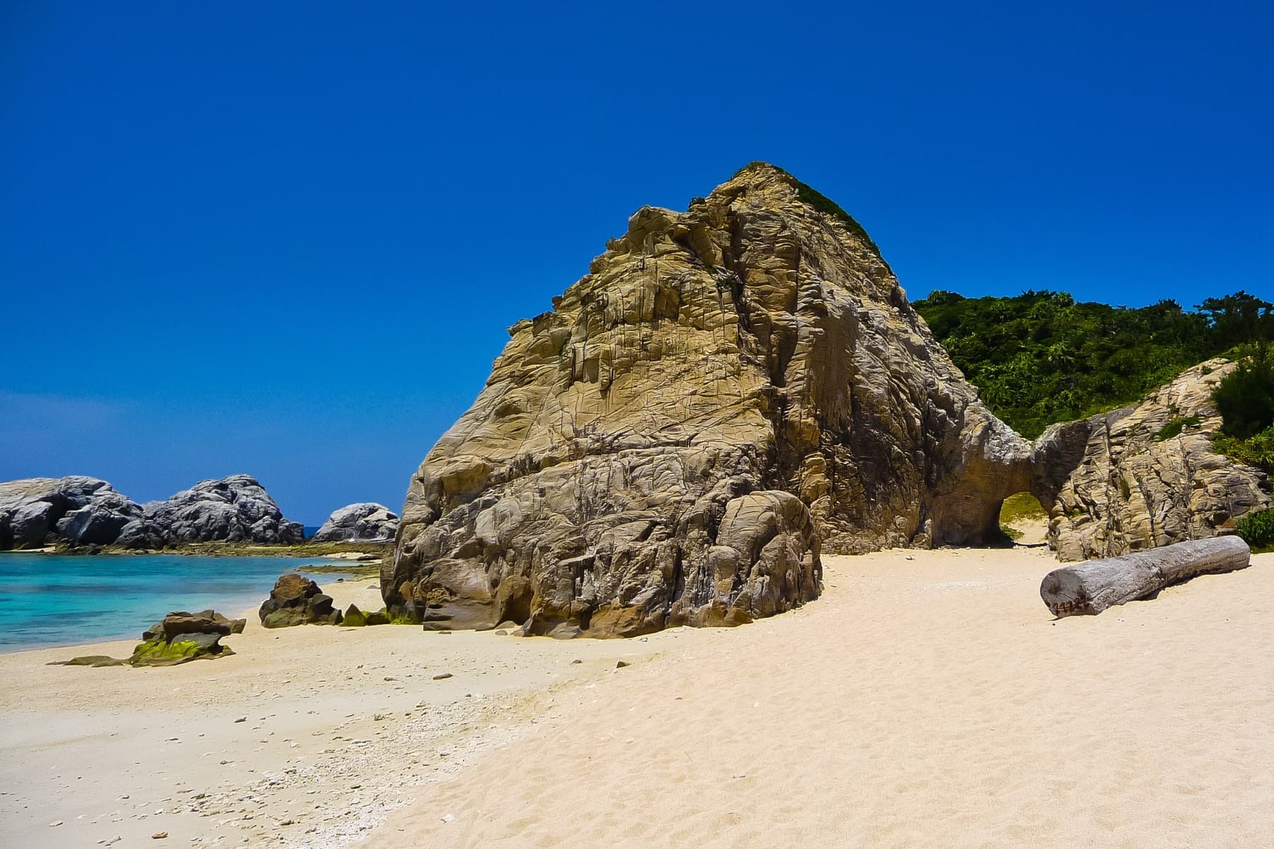 A tall weathered boulder stands on the sand at Aharen Beach on Tokashiki, and a bleached log lies beside it under a deep blue sky