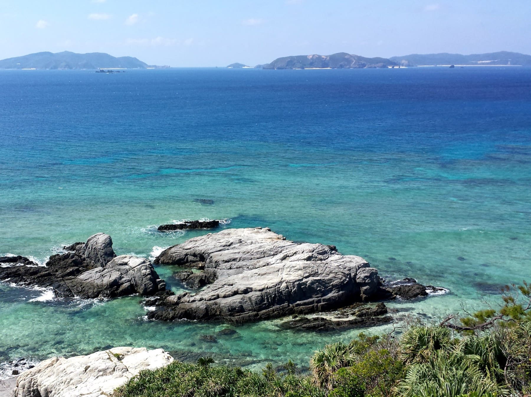 Grey rock slabs break the surface below the Aharen observation deck on Tokashiki, and the reef fades into deep blue toward the other Kerama islands
