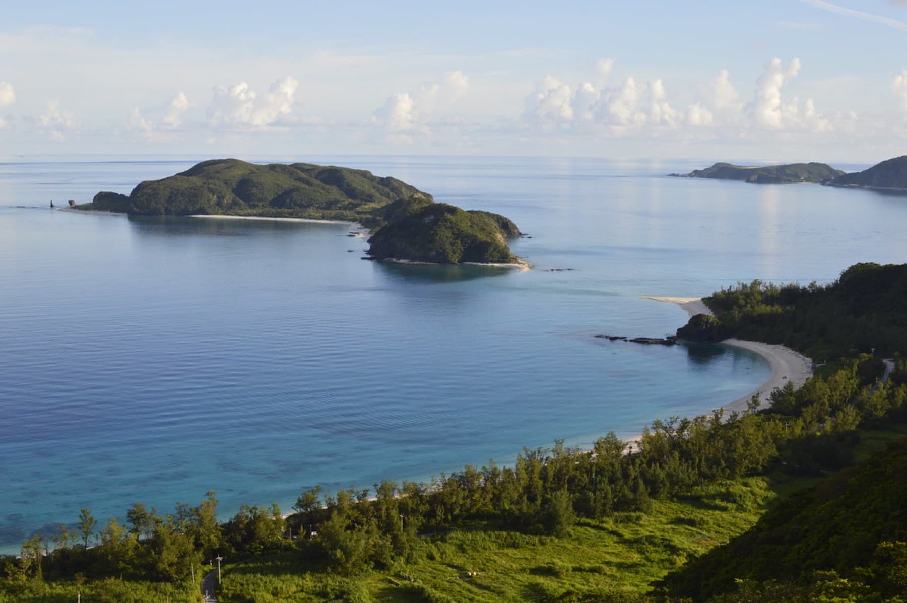 Soft morning light falls on a still blue sea seen from a green hill on Aka, where a white beach curves toward two small wooded islets