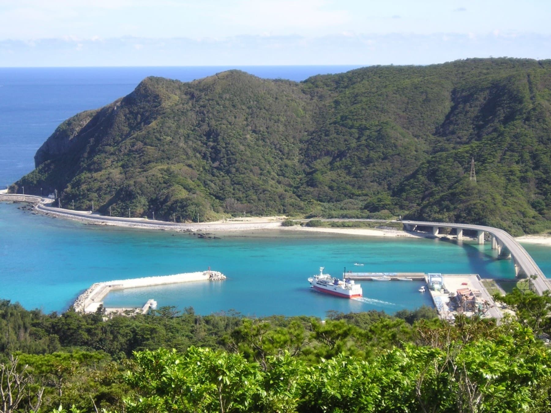 A ferry sits at Aka Port below a wooded hill on Geruma, and the curved Aka Ohashi bridge crosses the blue channel between the two islands