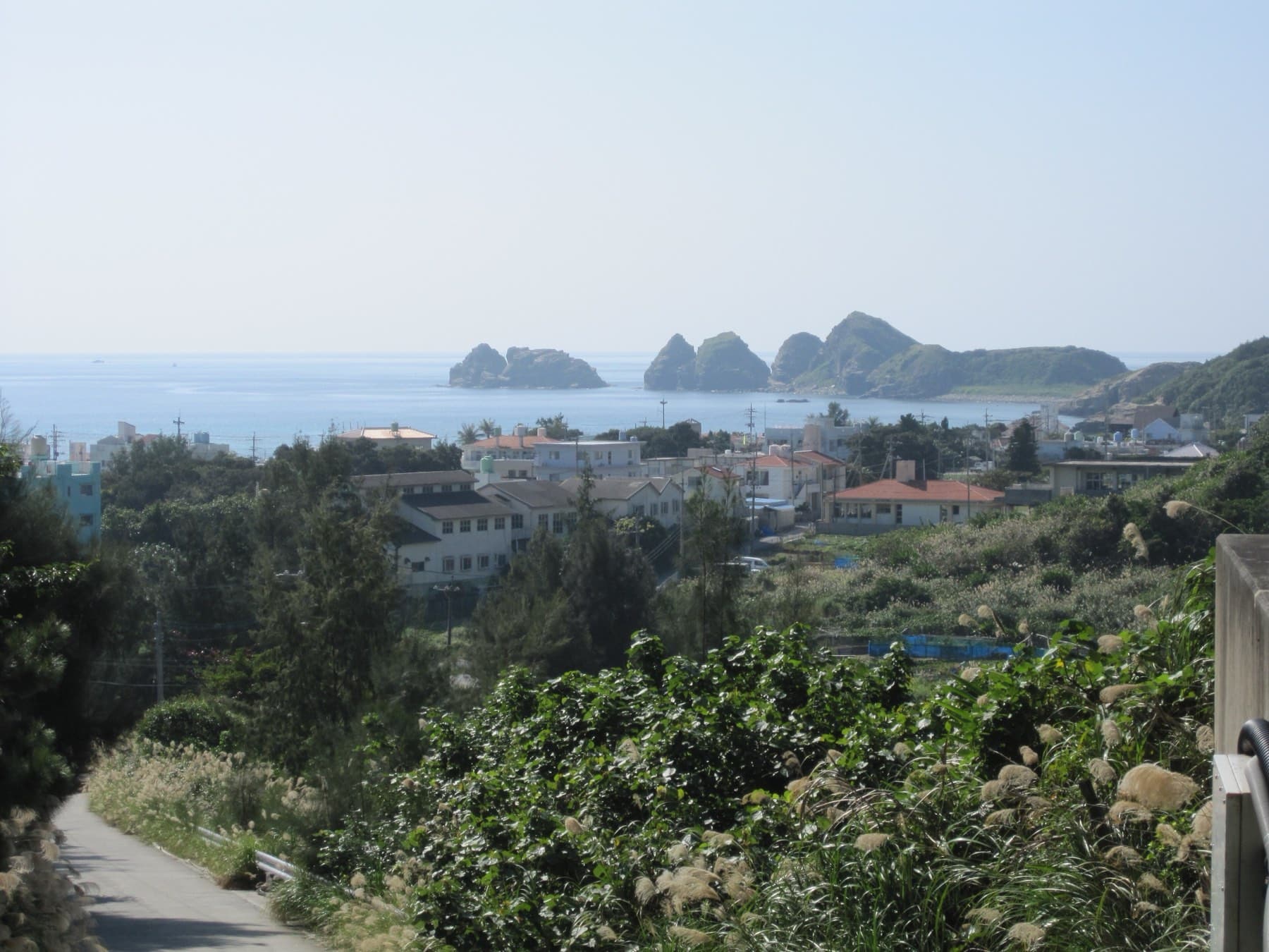 A narrow lane drops through scrub and silver grass toward the low roofs of Aka village, with rocky islets standing in the sea beyond under a hazy sky.