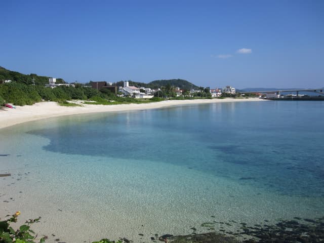 Clear shallow water curves along the village beach at Aka, where low houses sit behind the sand and the Aka Ohashi crosses toward Geruma at the far end