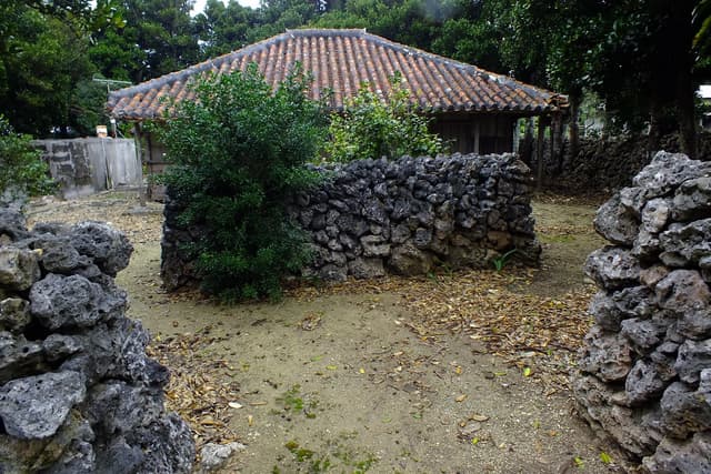 An old wooden house under a red-tile roof stands behind a dry coral stone wall and a gap-toothed gateway in the shade of trees on Hateruma