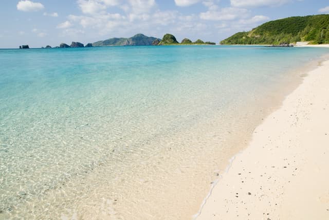 Clear shallow water laps the white sand of Ama Beach on Zamami as the rocky outlines of small uninhabited islets line the horizon to the south