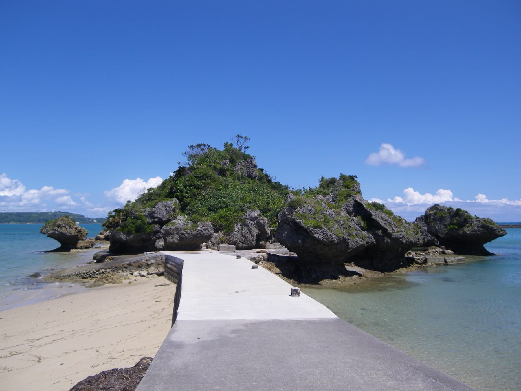 A concrete walkway crosses the sand to the rocky green islet of Amamichu off Hamahiga, with clear shallow water on both sides under a deep blue sky