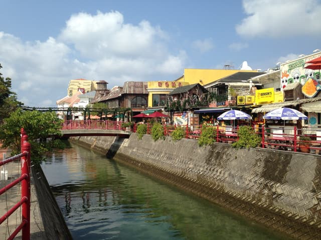 A narrow canal runs between stone walls through American Village, with a red footbridge, cafe umbrellas and painted shopfronts along the far bank