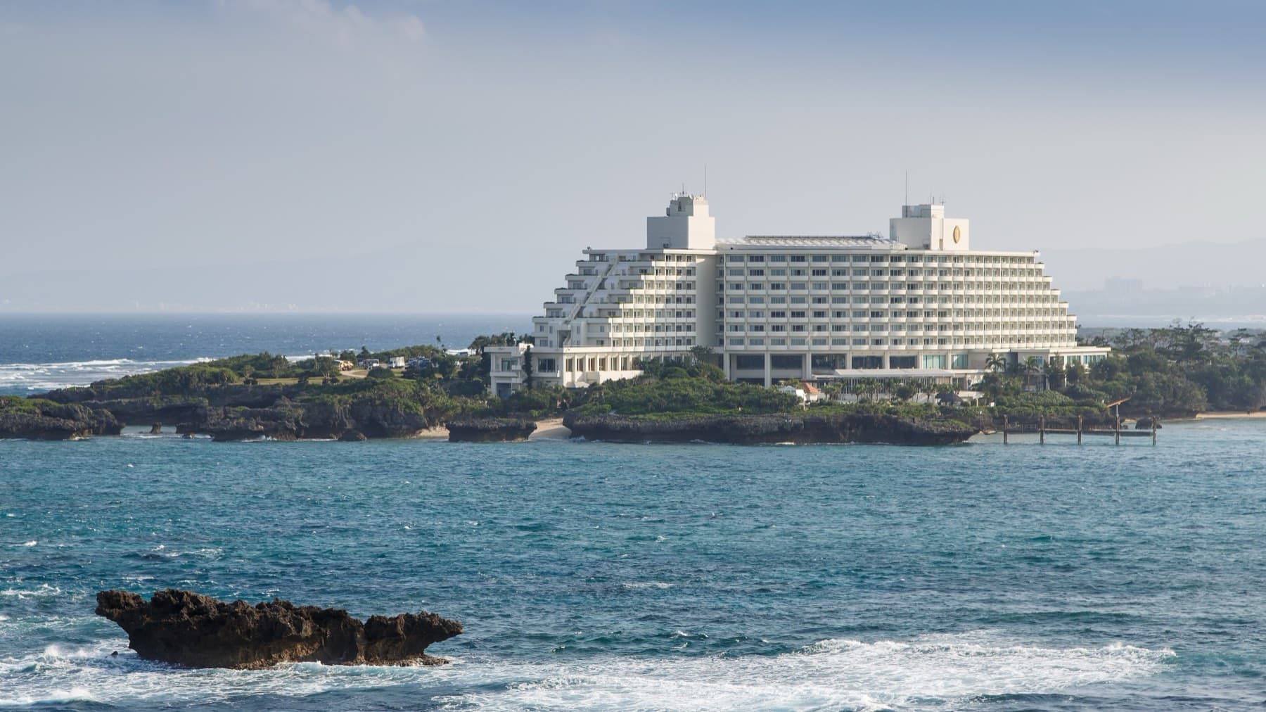 The stepped white tower of the ANA InterContinental Manza Beach Resort fills the tip of a low wooded point, with blue water and a dark reef rock in front