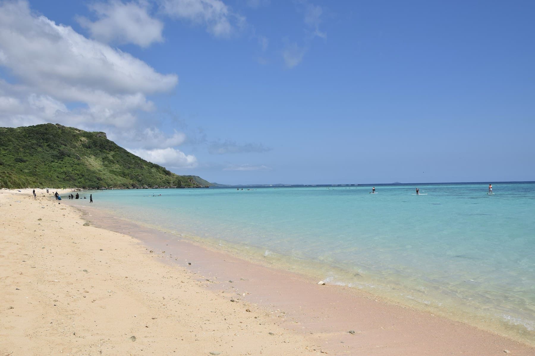 Clear shallows over the reef lap a long strip of pale sand at Aragusuku Beach as a green headland closes the far end of the bay.