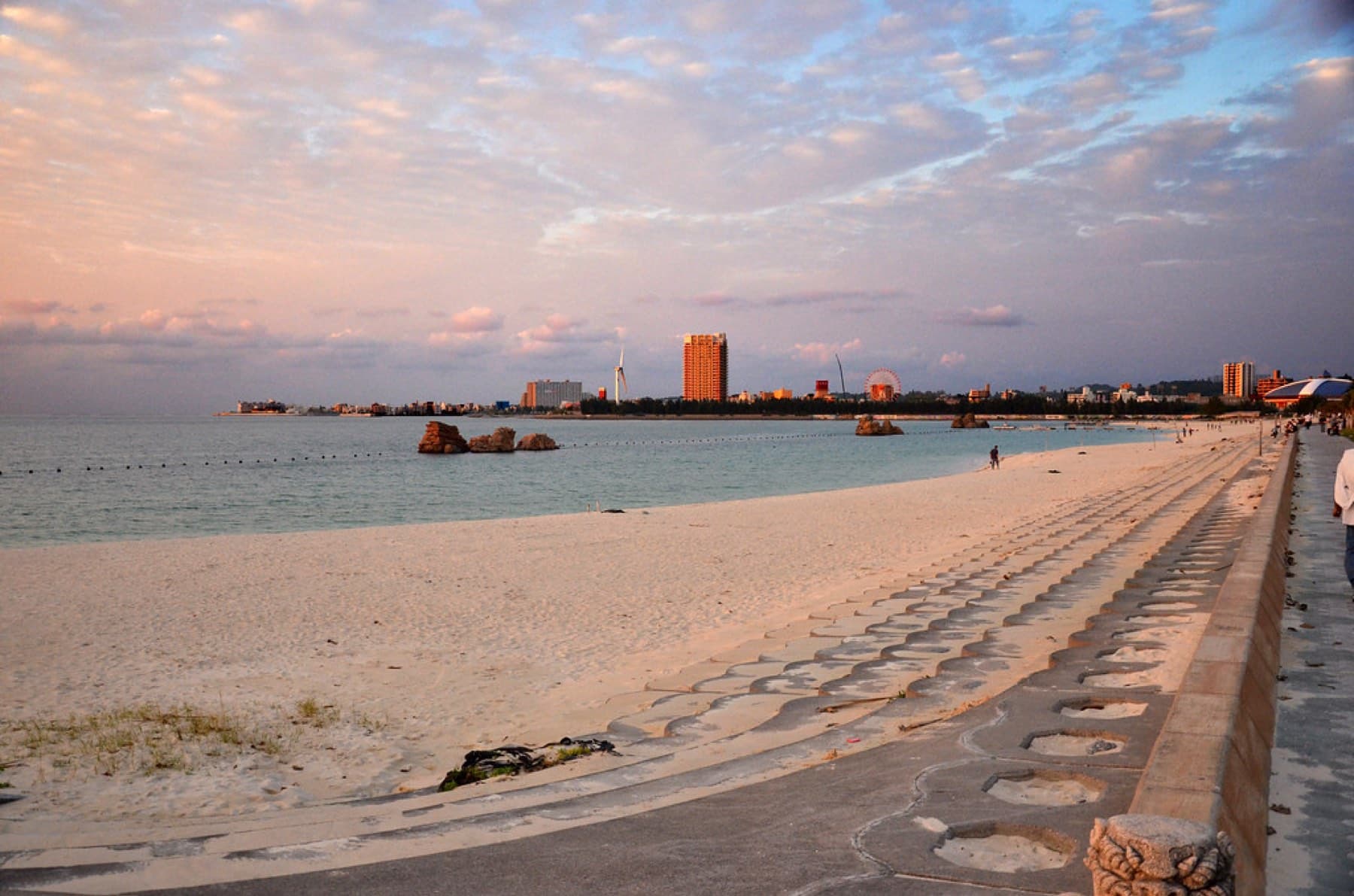 Pink evening cloud hangs over the long curve of Araha Beach, where stepped concrete edges the sand and rocks stand in the calm water
