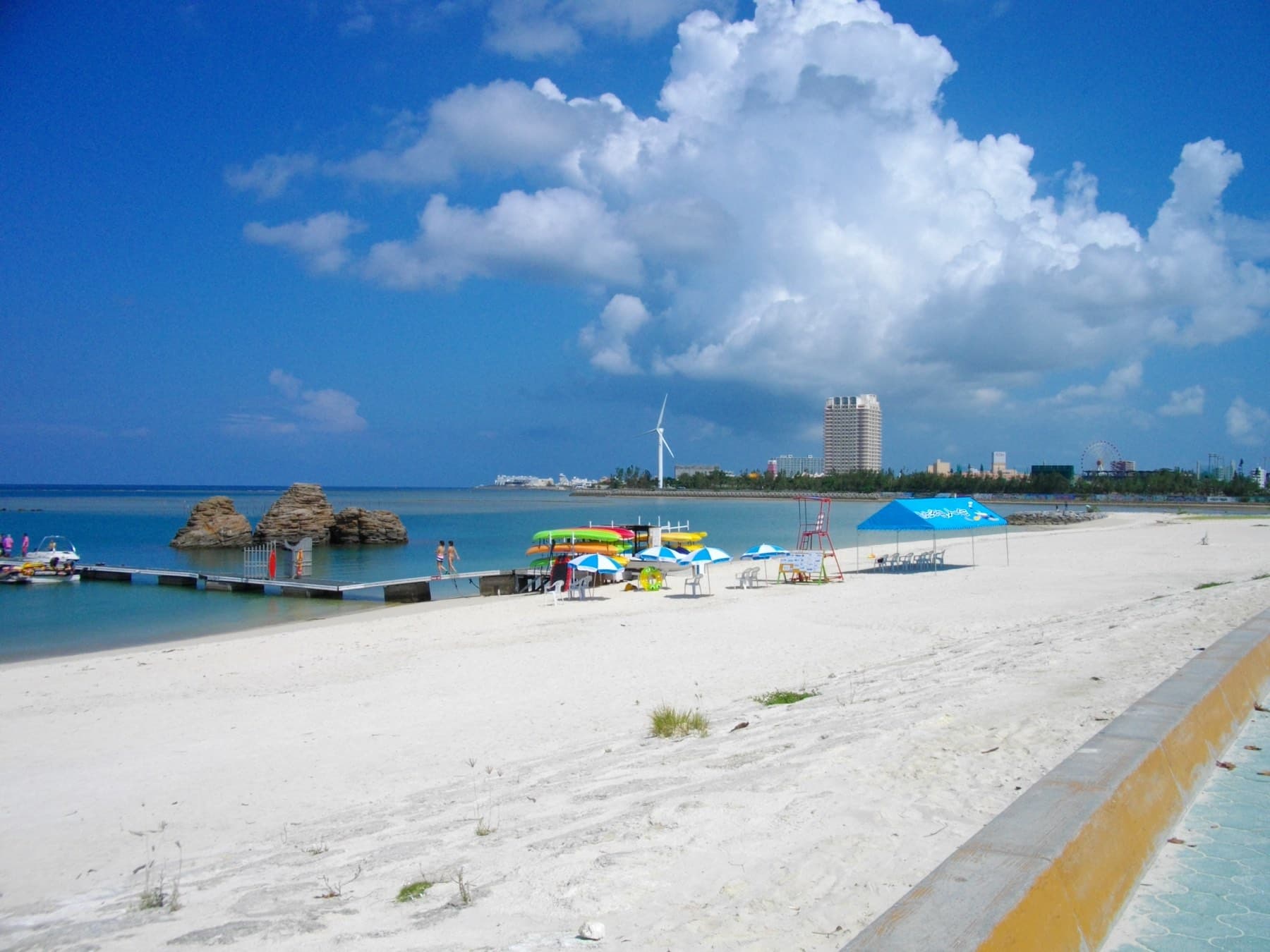 Wide white sand runs along Araha Beach in Chatan toward a jetty and offshore rocks, with beach umbrellas and a tall summer cloud over the city
