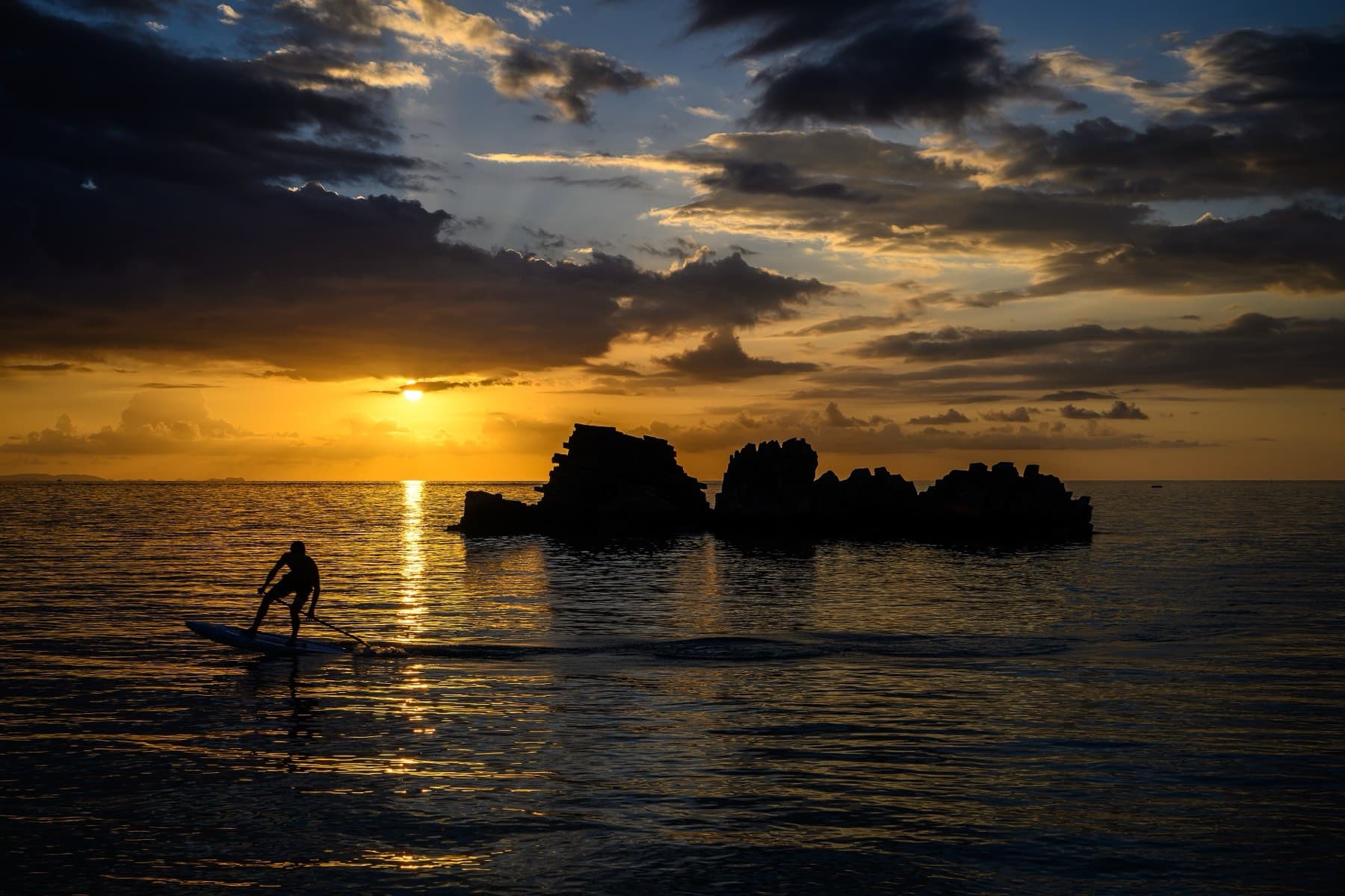 A paddleboarder glides in silhouette across golden water off Araha Beach as the sun sets behind dark rocks and streaks of cloud over the East China Sea