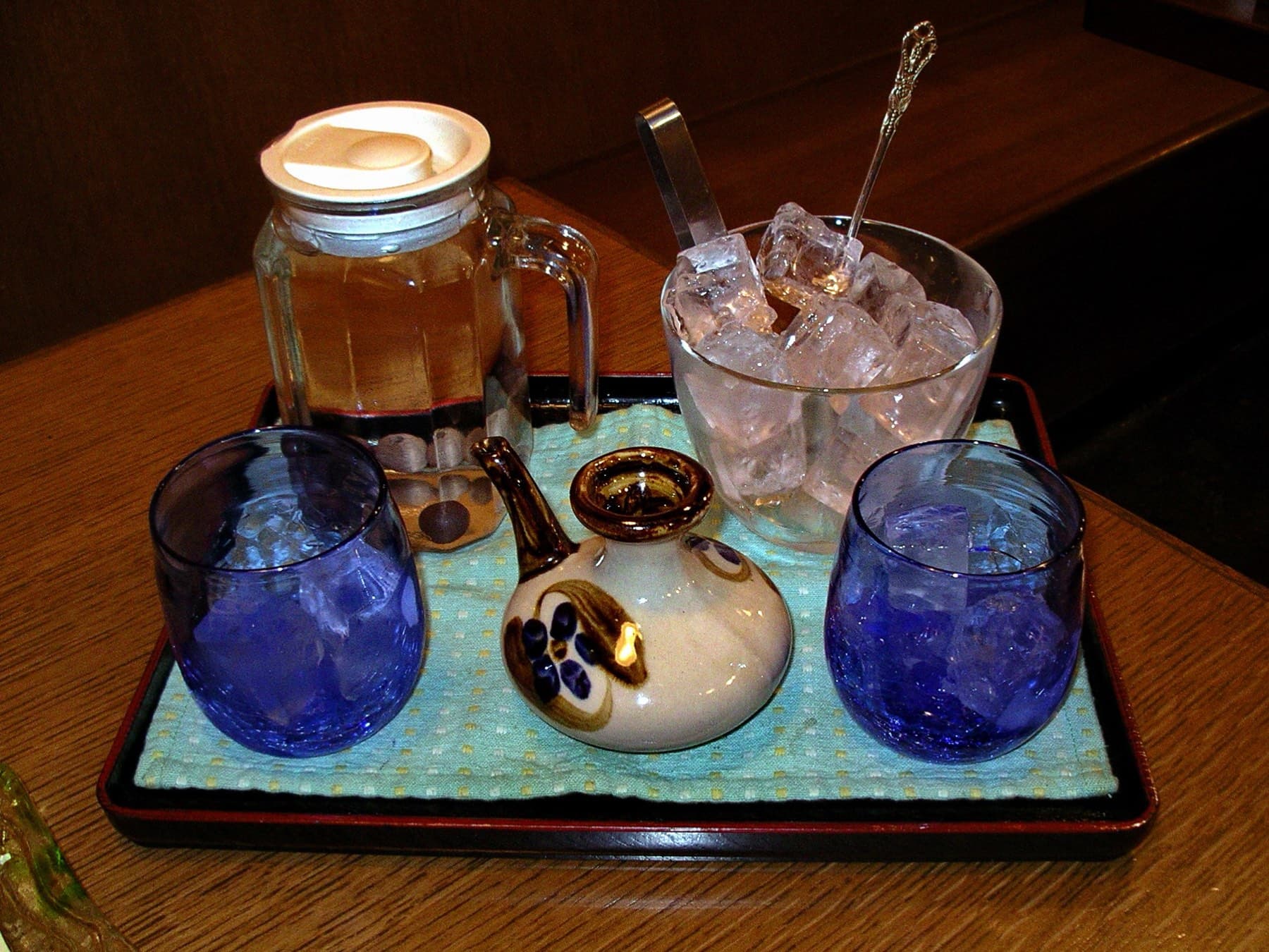 Two blue Ryukyu glass tumblers filled with ice flank a painted karakara flask of awamori on a lacquer tray with a water jug and a bowl of ice