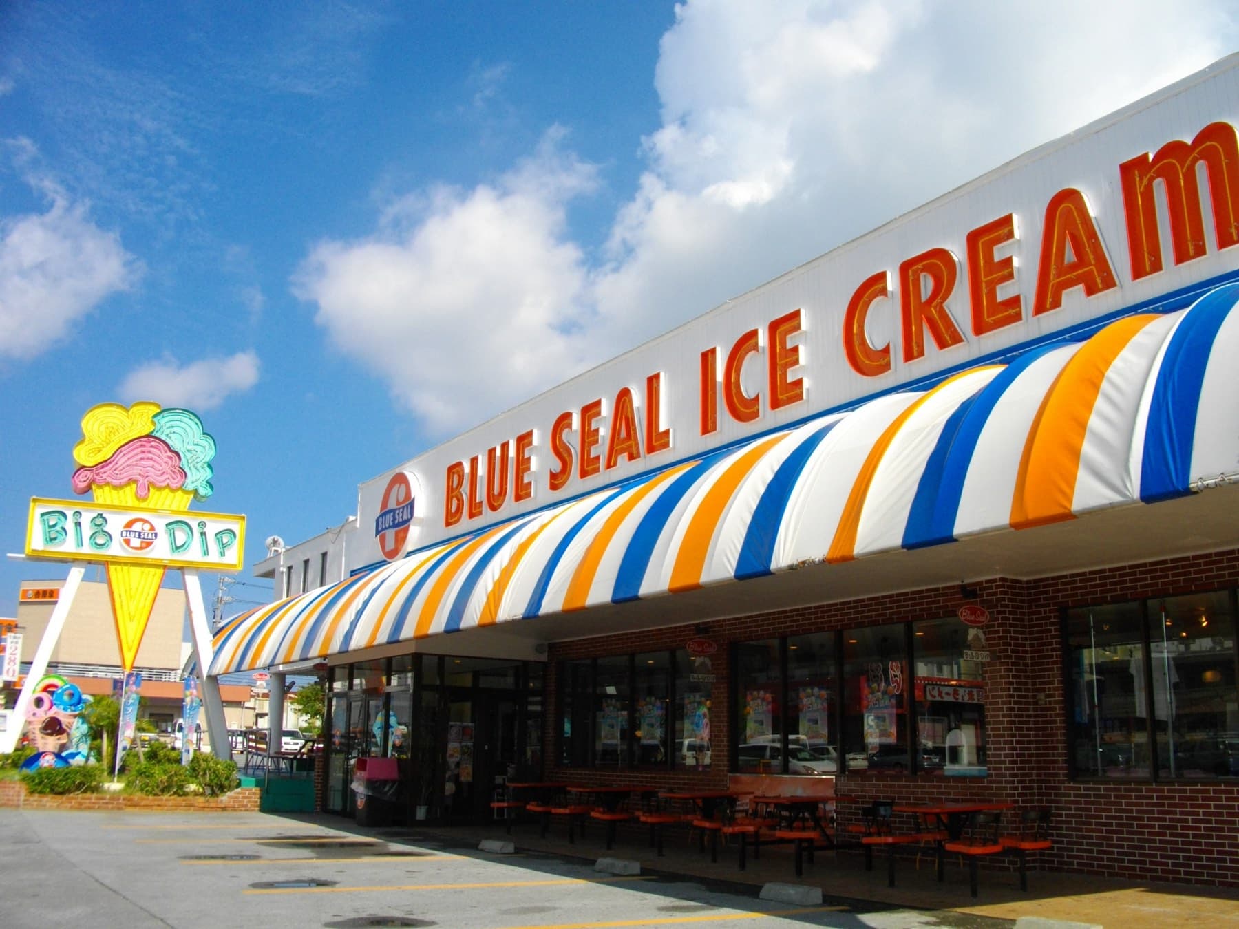 An orange, white and blue striped awning runs along the Blue Seal Ice Cream shop in Makiminato beside its tall Big Dip cone sign under a blue sky