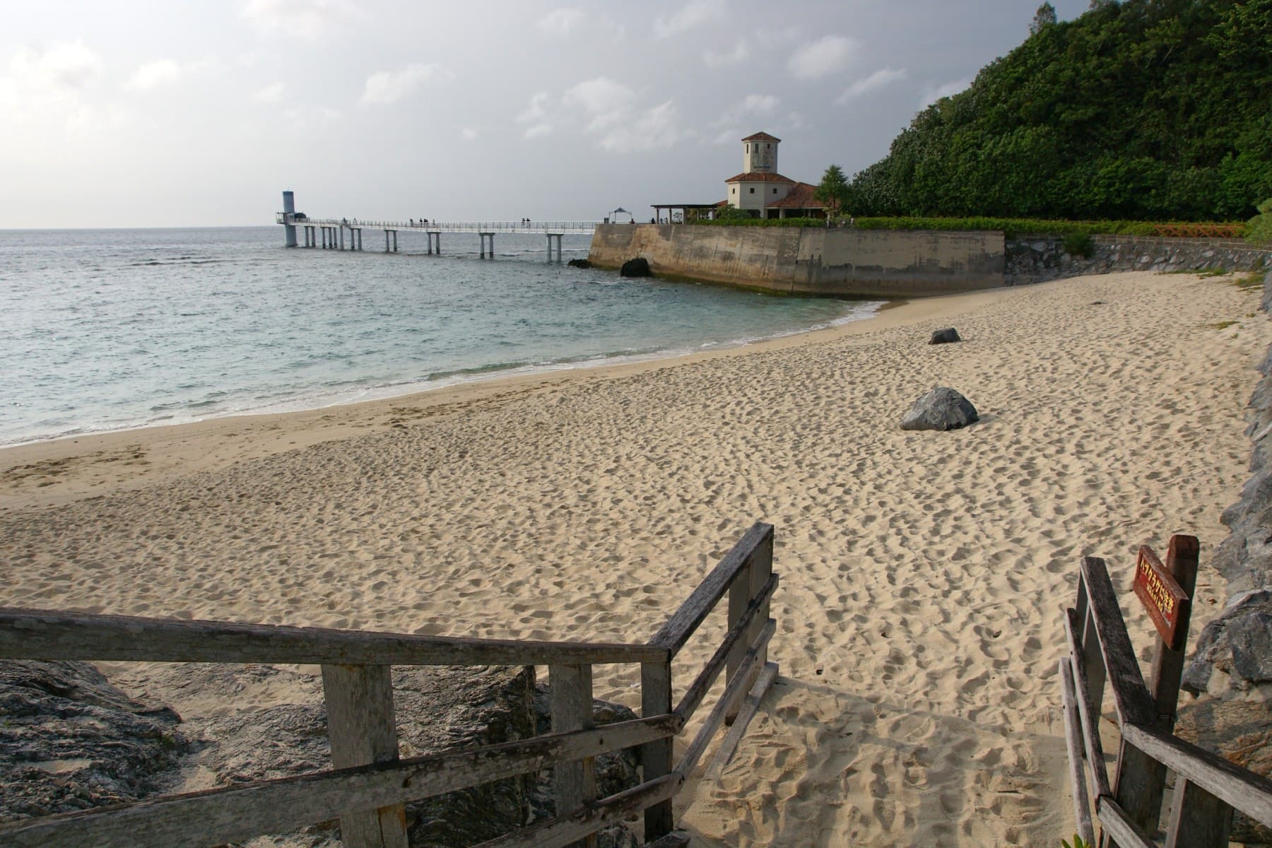 A wooden stair leads down to the sand on the Busena peninsula in Nago, and the long pier runs out over grey-green water to the underwater observatory