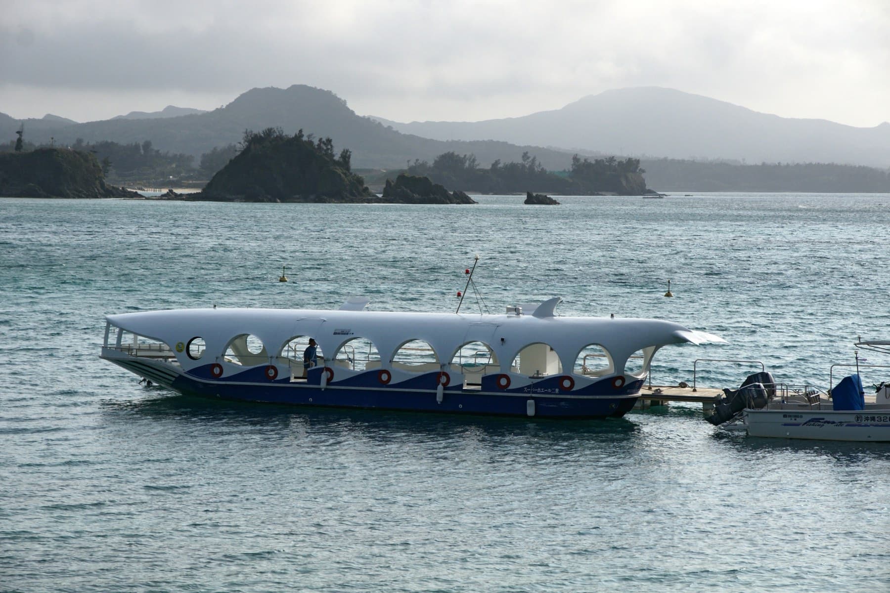 A whale-shaped glass-bottom boat with round portholes waits on calm water at Busena Marine Park, with rocky islets and the hills of Nago behind