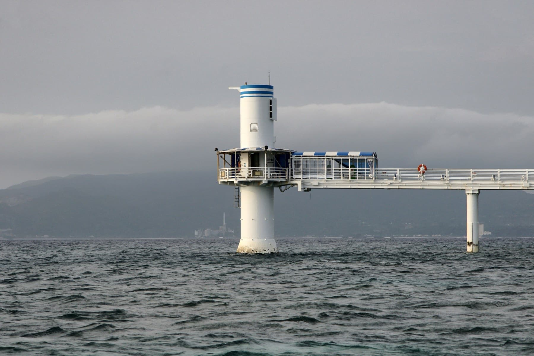 The white tower of the Busena underwater observatory stands at the end of its long pier in grey water off Nago as cloud rolls over the hills