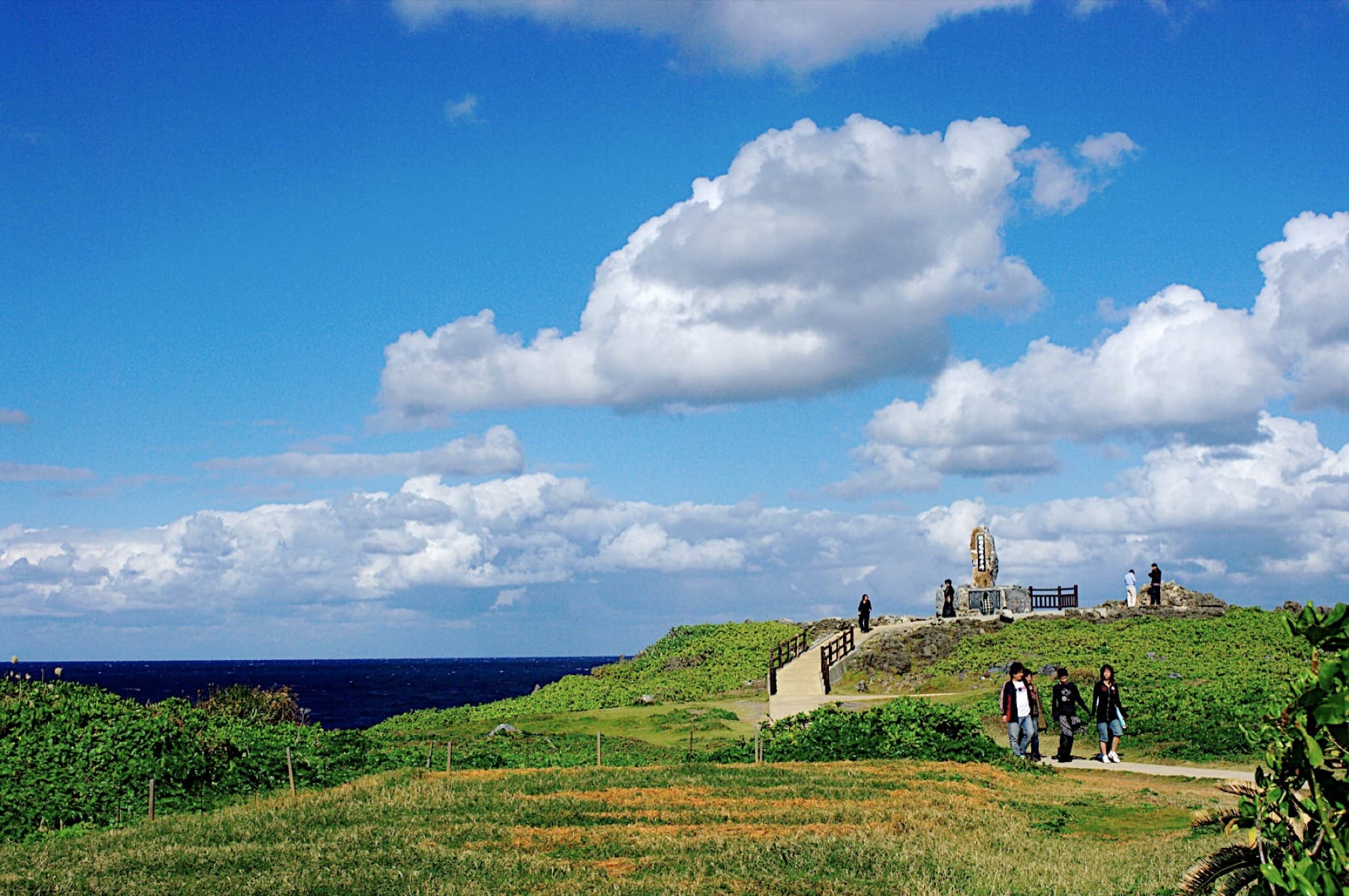Visitors walk a path across green turf toward the stone monument on Cape Hedo as white cumulus clouds pile up over the dark sea