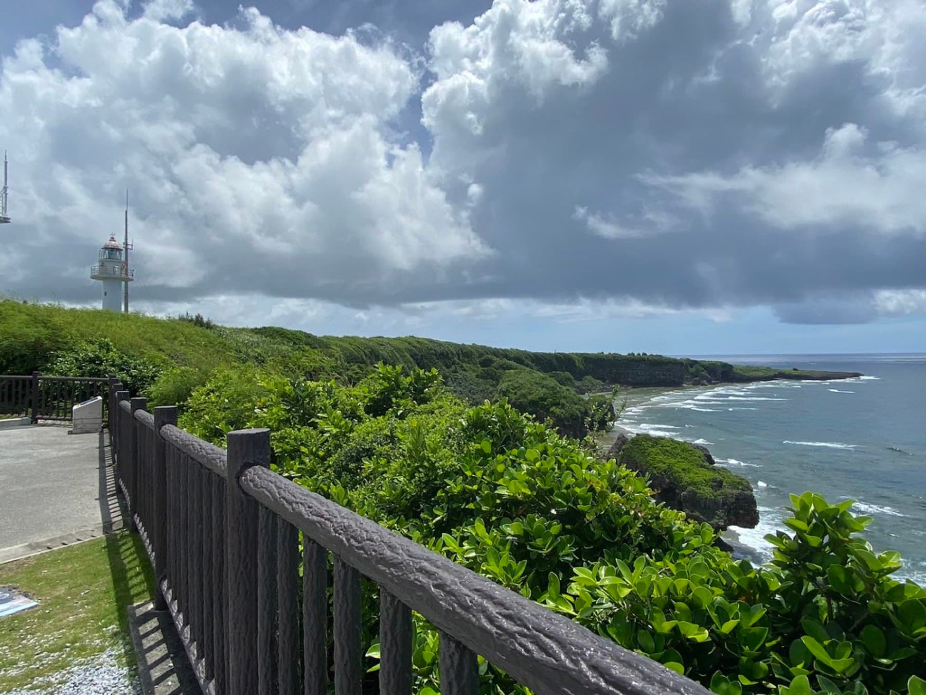 A wooden railing at Cape Kyan looks along green clifftops to a curving reef coast, with the lighthouse on the left and heavy summer clouds overhead