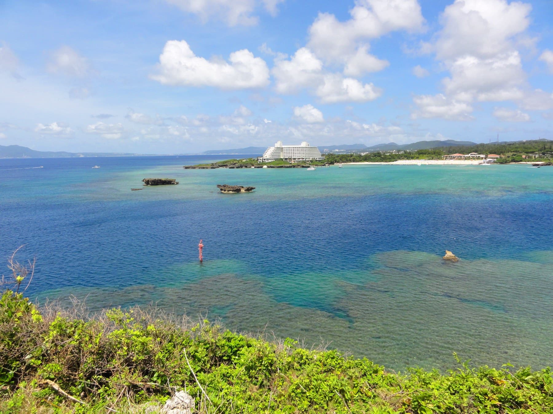 Green scrub on Cape Manzamo frames a view over Manza Bay, where reef shows through the clear water and the white ANA InterContinental resort sits on the far point