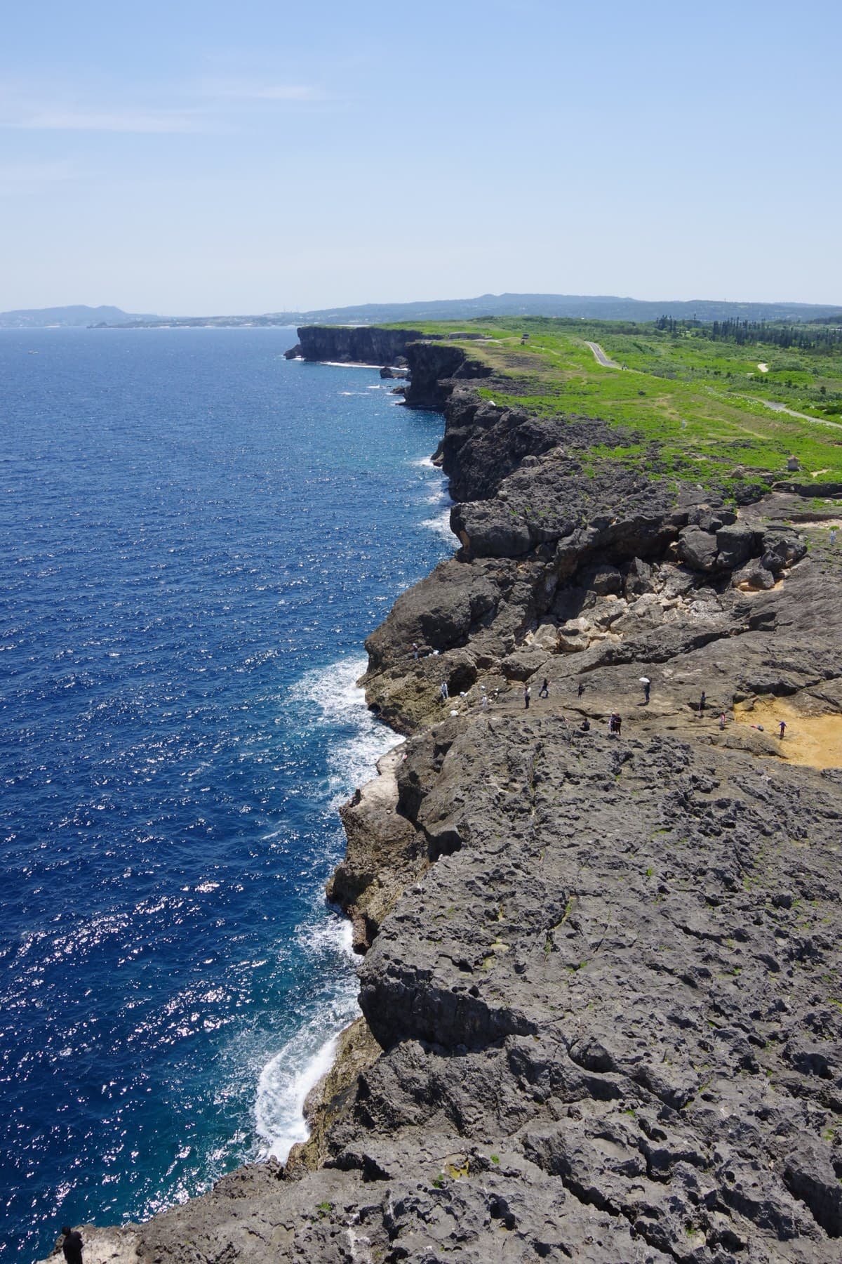 Dark limestone cliffs run for kilometres along the deep blue sea at Cape Zanpa in Yomitan, seen from the top of the lighthouse with green fields inland