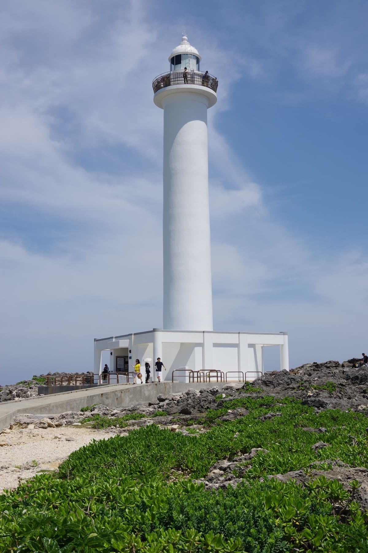 The tall white Zanpa Misaki lighthouse rises from a low white building and green coastal plants under a blue sky with drifting clouds