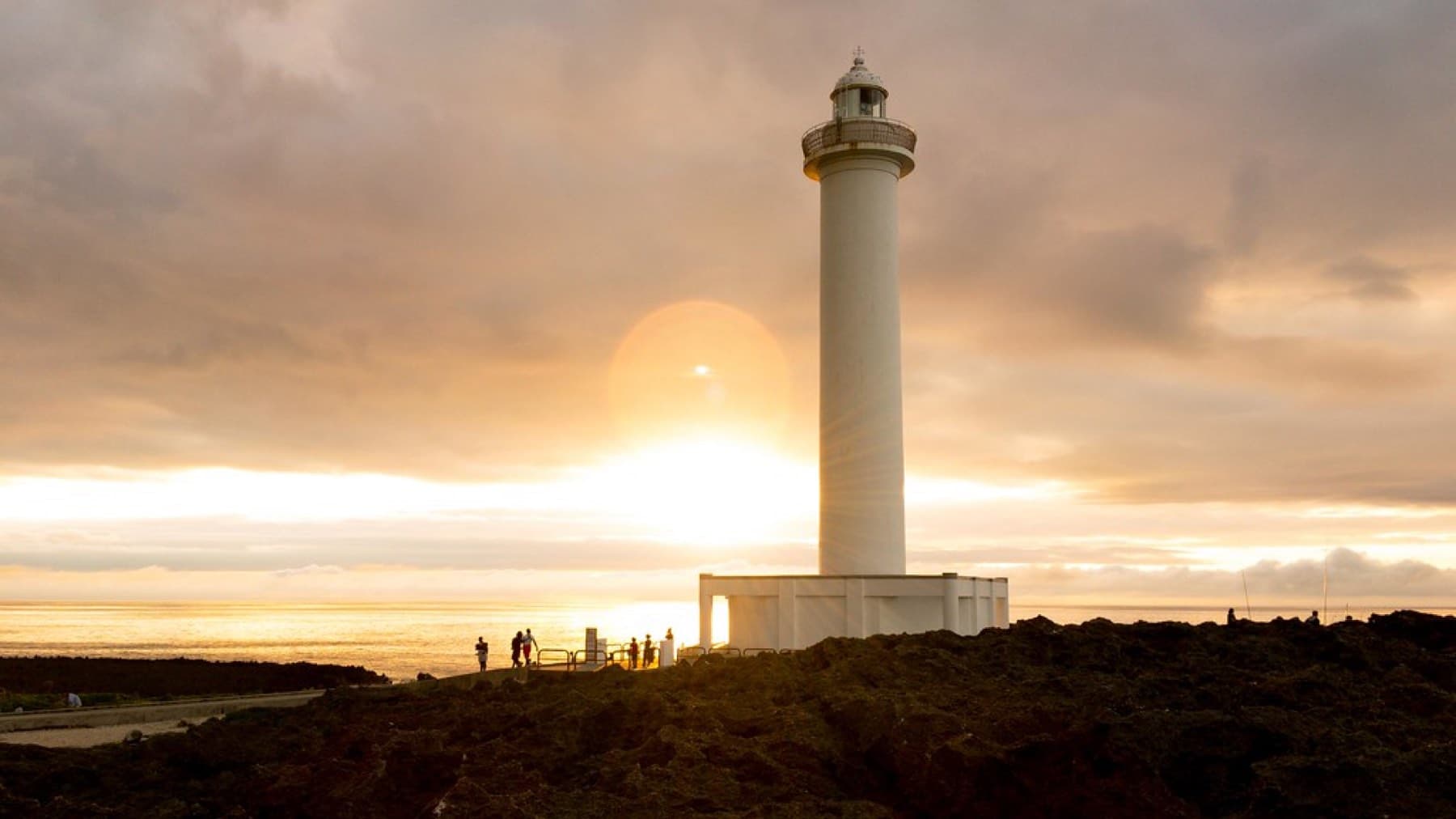 The sun sets behind the Zanpa lighthouse over the East China Sea while a few people stand on the dark rocks of the cape in golden light