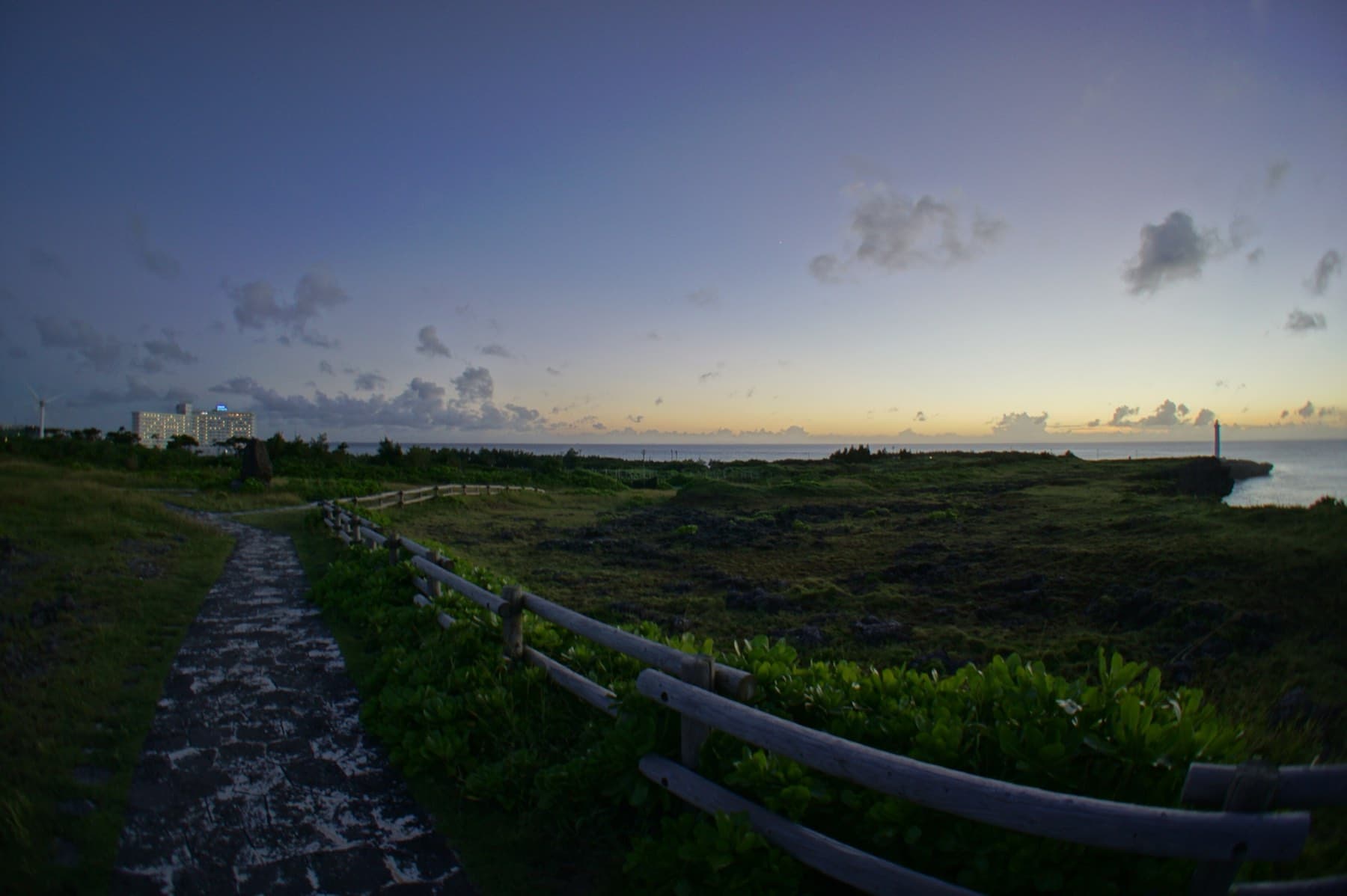 A stone path with a wooden rail crosses the low scrub of Cape Zanpa at dusk, with the lighthouse on the far headland and a band of orange light fading over the sea.