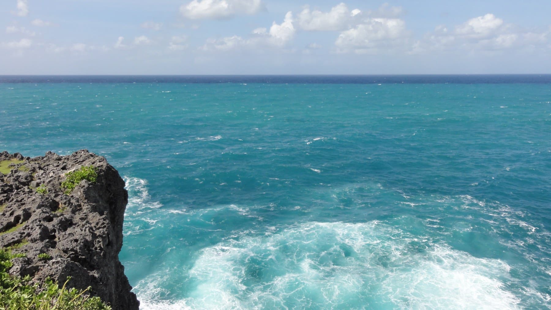 Turquoise water boils white against the dark limestone cliffs of Cape Zanpa, with the open East China Sea stretching to a horizon of small clouds.