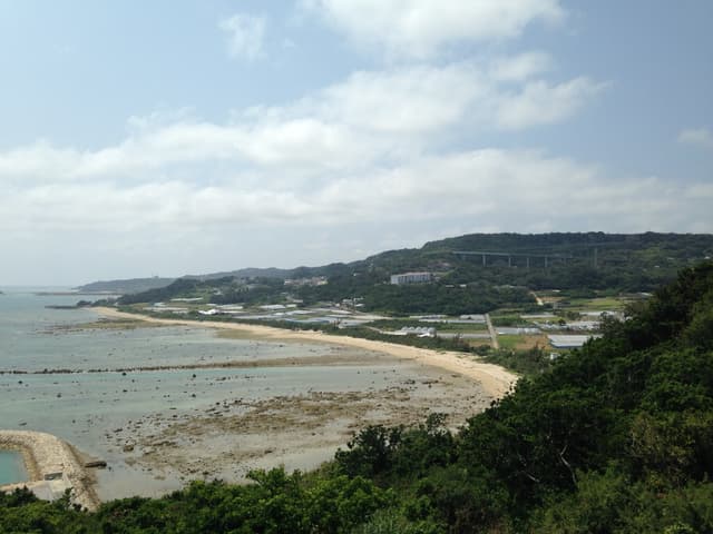 A long curve of sand and a wide reef flat at low tide run below green hills on the Chinen coast, seen from Chinen Misaki Park