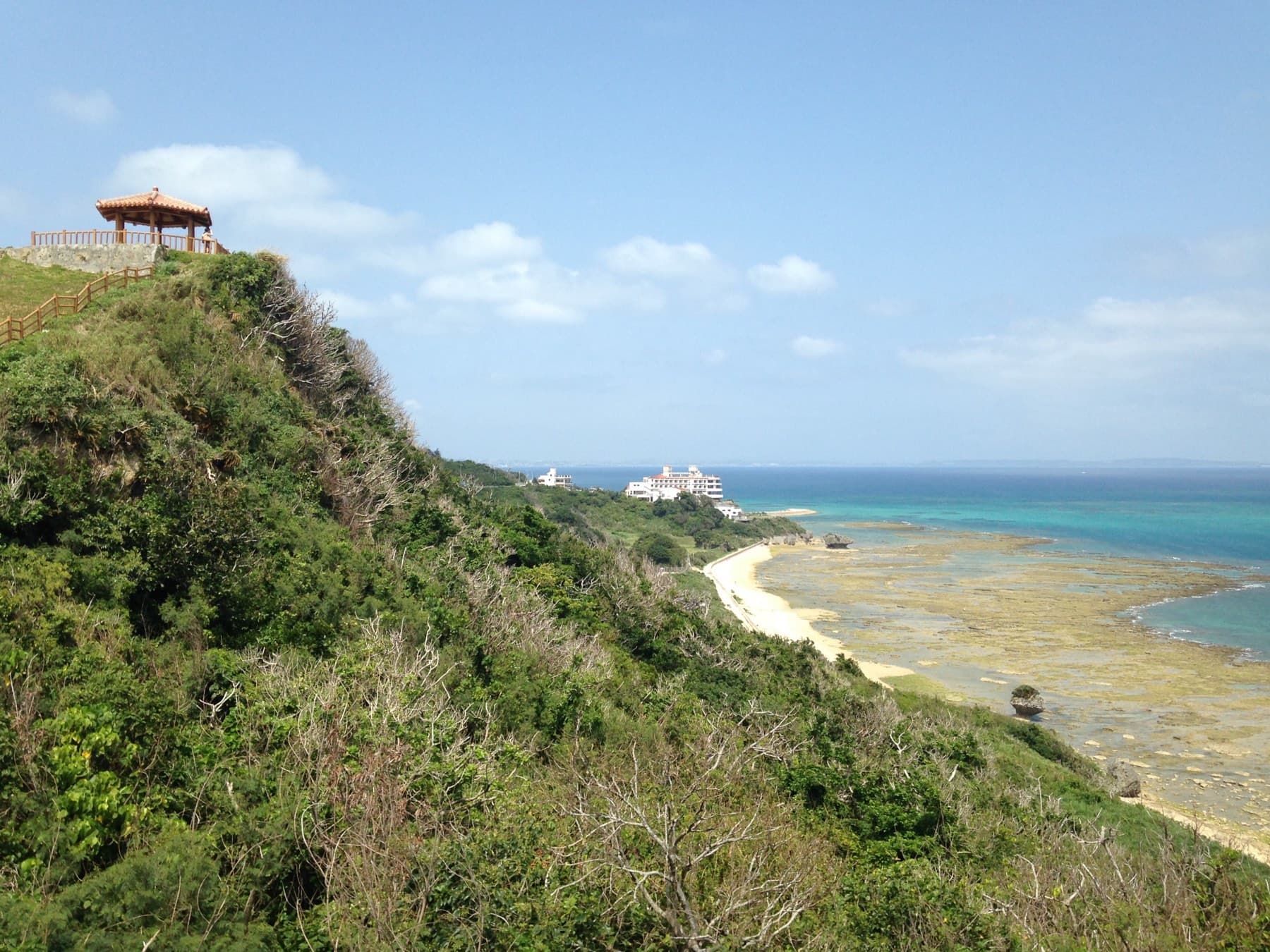 A lookout pavilion crowns a steep green bluff at Chinen Misaki Park, with a sandy beach and wide reef flat curving away below