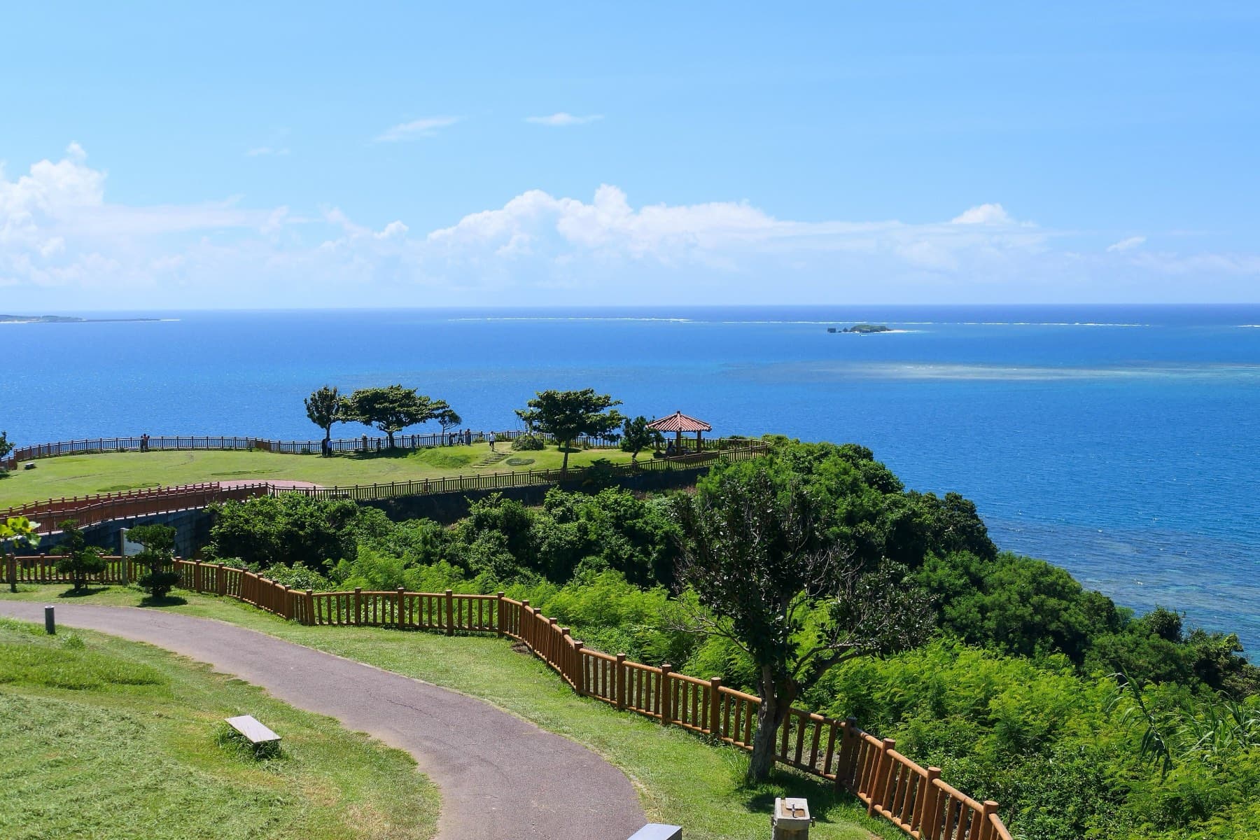 A path curves down through Chinen Misaki Park to a grassy headland with a red-roofed pavilion, and Kudaka lies low on the deep blue sea beyond