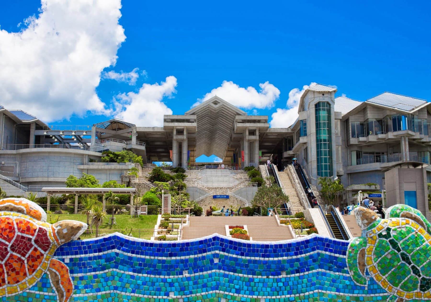 Mosaic sea turtles frame the terraced entrance of Churaumi Aquarium in Ocean Expo Park under a bright summer sky with tall white clouds