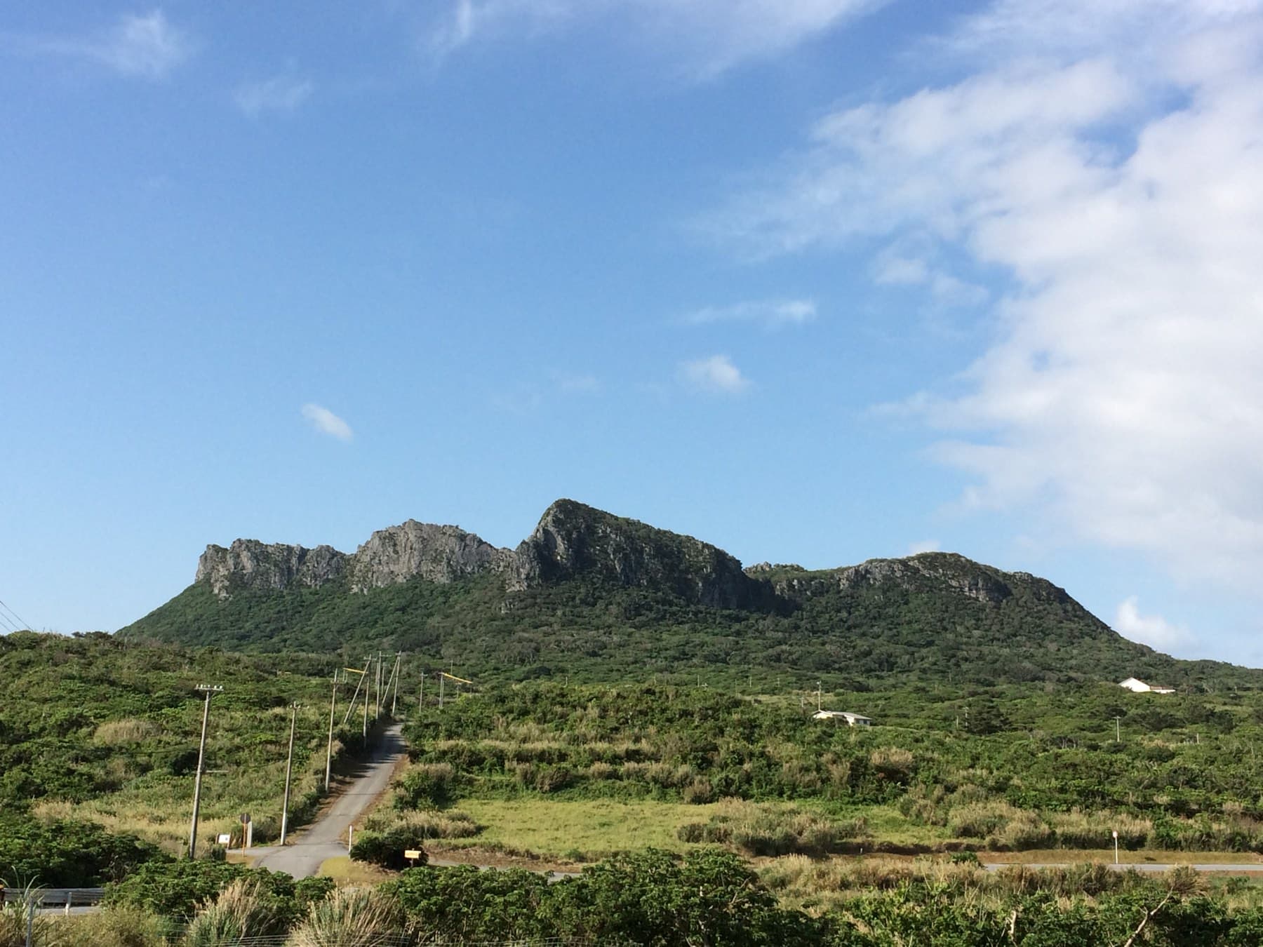 The jagged limestone crown of Asumui rises above green scrubland, and a narrow road runs toward it from Cape Hedo under a wide blue sky