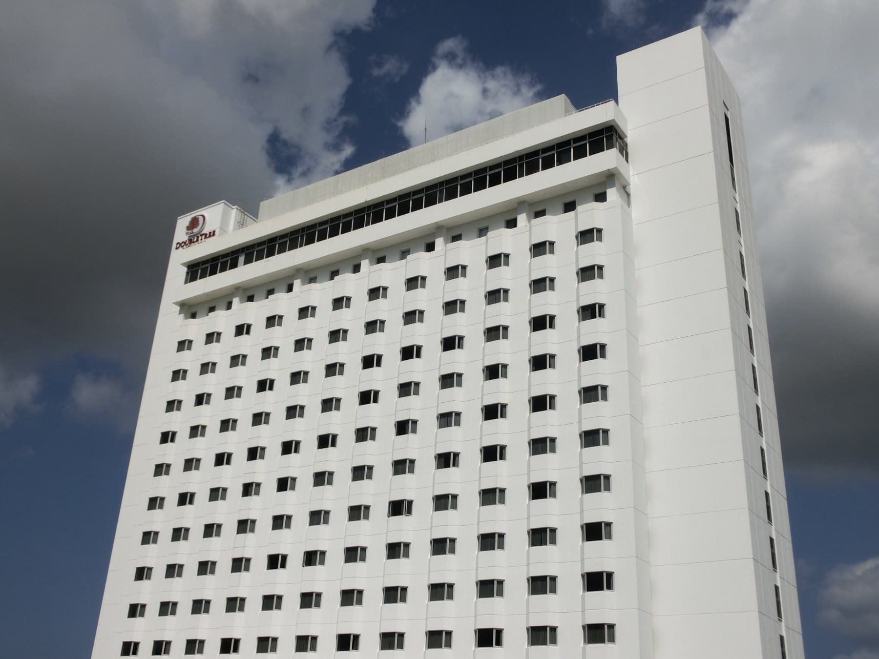 The white tower of DoubleTree by Hilton Naha Shuri Castle carries the hotel's logo near its roof under a cloudy sky.