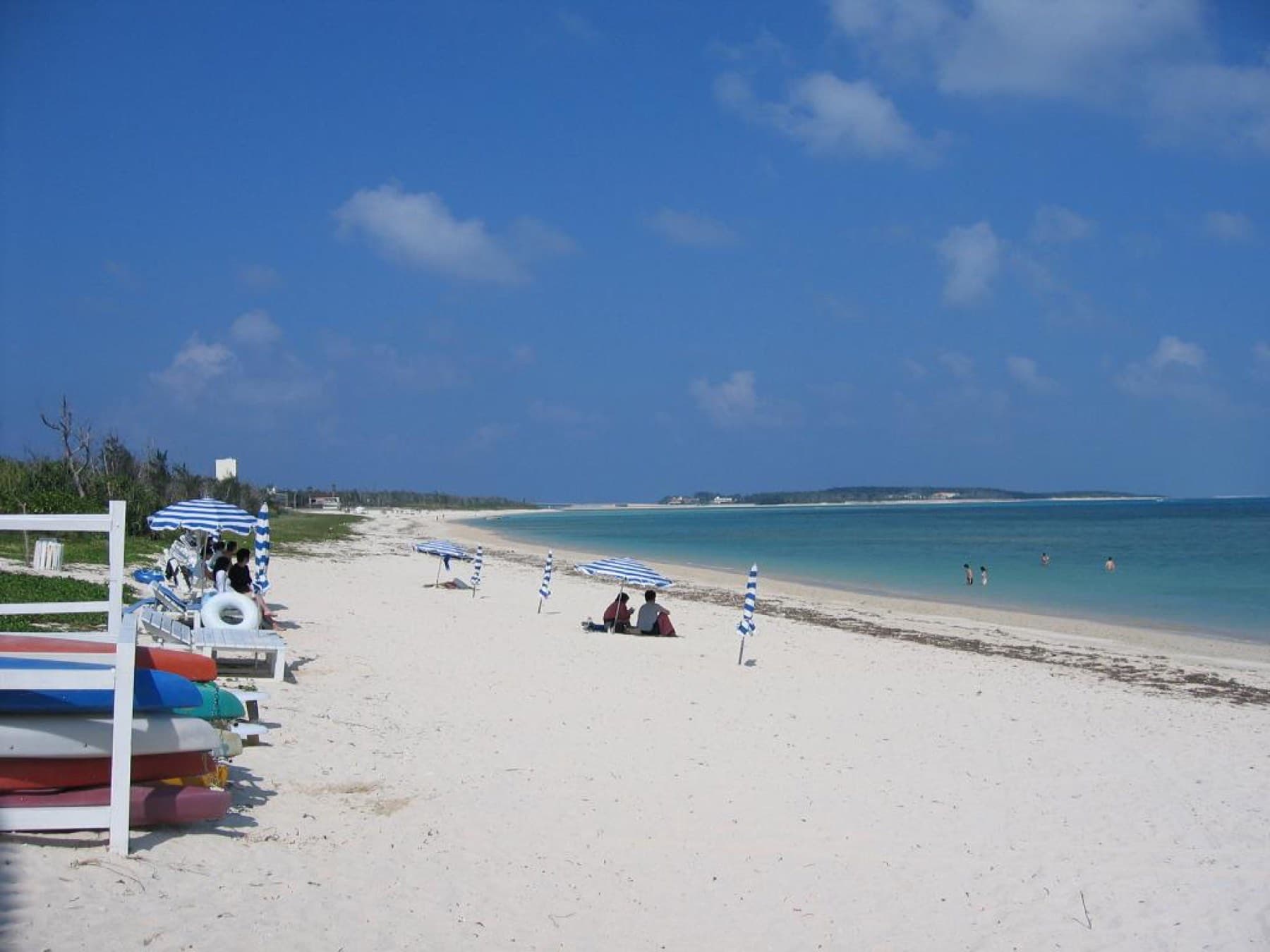 Striped parasols and stacked kayaks line the white sand of Eef Beach on Kume-jima as the shallow bay curves toward Ou-jima under a deep blue sky