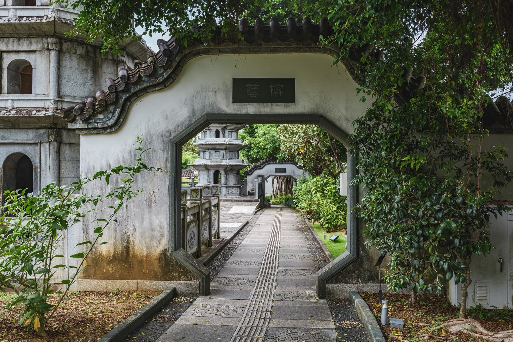An eight-sided gateway in a white wall frames a paved path leading past stone balustrades to a pale stone pagoda in the Fukushuen garden