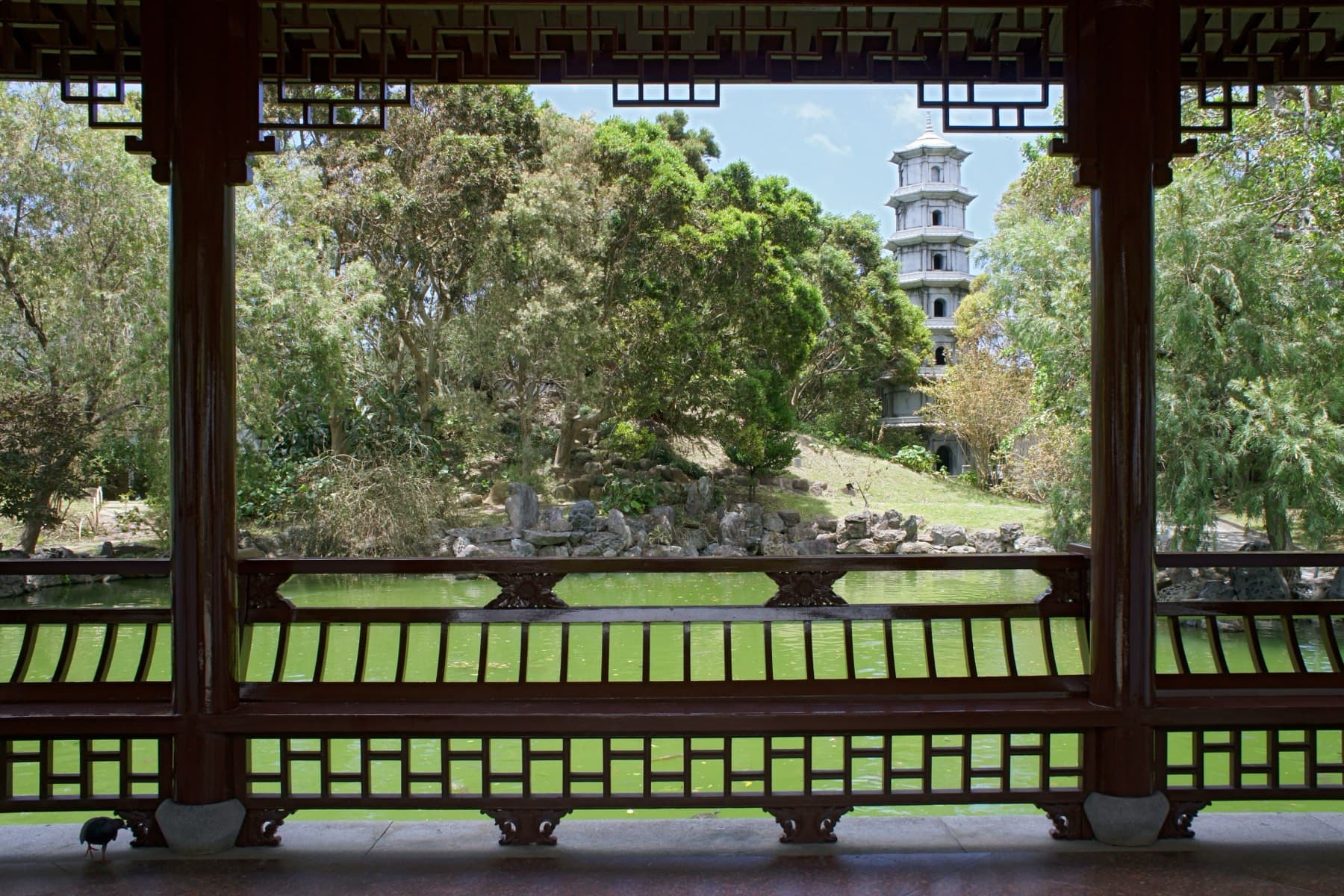 Lacquered posts and a carved green railing frame a view across the Fukushuen pond to a white stone pagoda among the trees