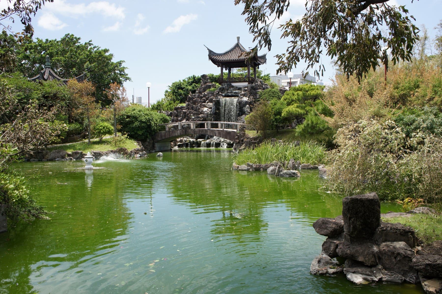 A green pond fills the foreground at Fukushuen in Naha, and a Chinese pavilion stands on a rock hill above a waterfall at the far end