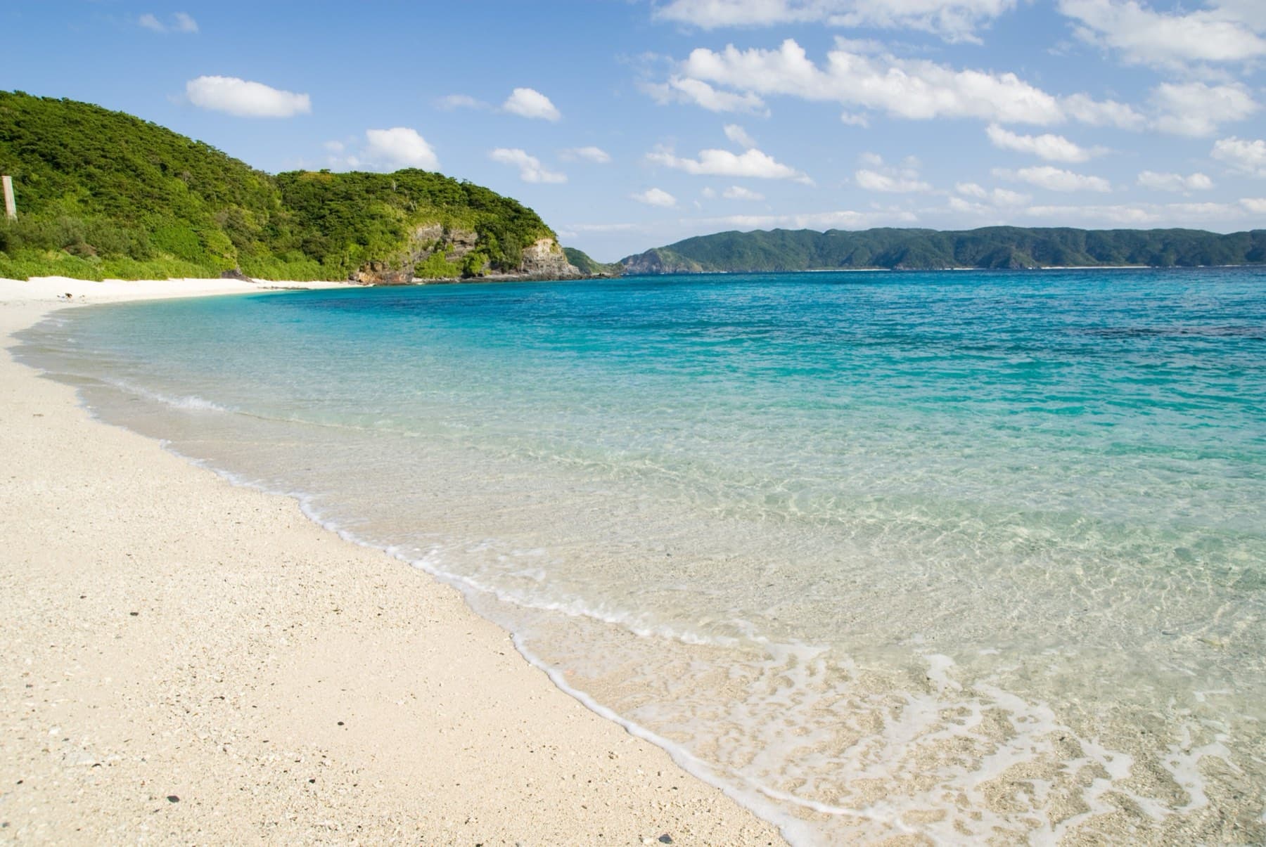 White sand curving along Furuzamami Beach on Zamami, with clear shallows over the reef and green headlands behind