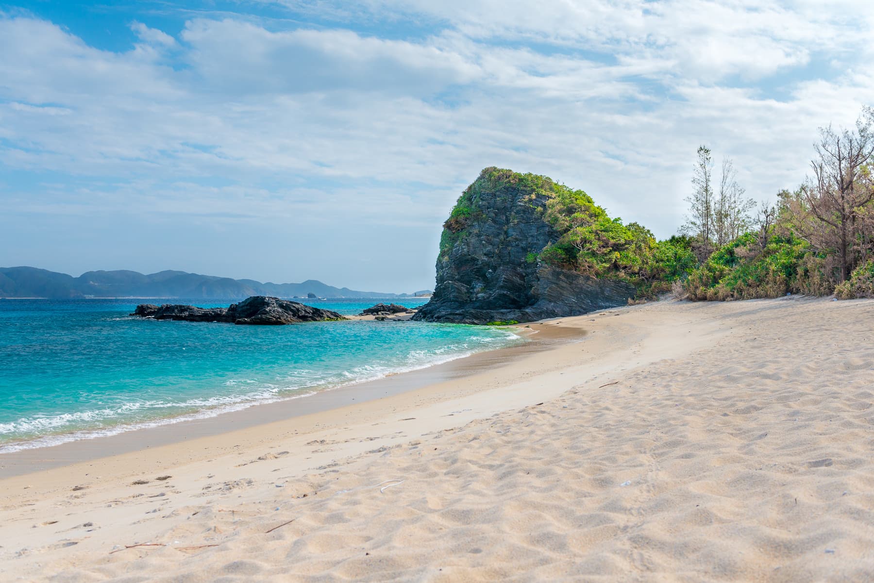 Fine sand slopes into bright blue water at Furuzamami Beach, where a rock headland covered in scrub closes off the bay under thin cloud