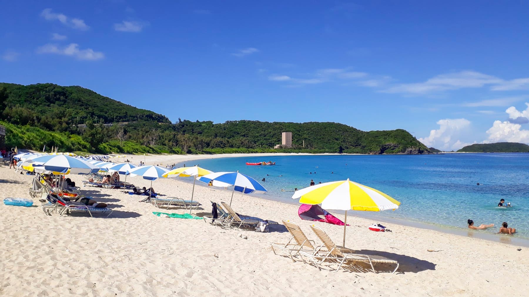 Yellow and blue parasols shade loungers on the white sand of Furuzamami Beach in July while swimmers float in the calm water by the reef