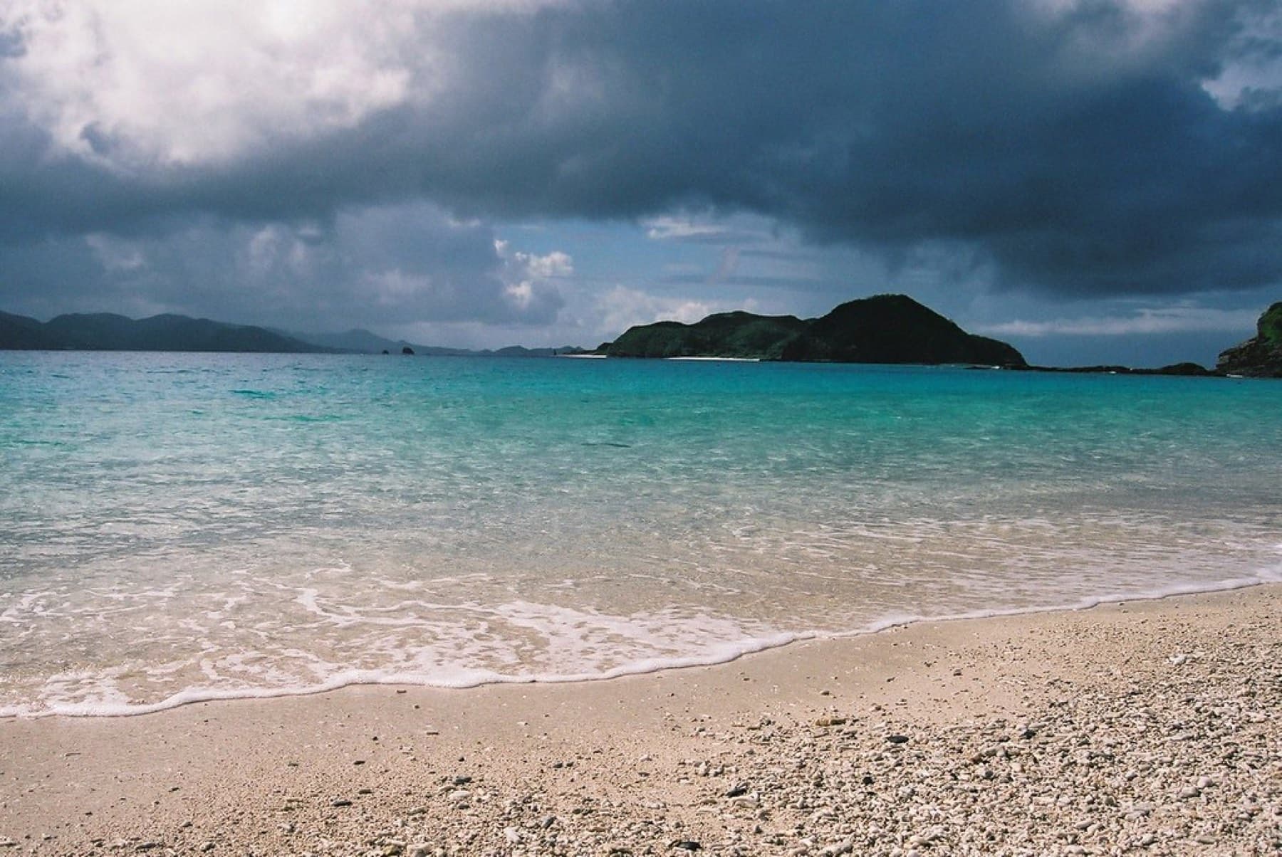 Dark storm clouds gather over the small islets off Furuzamami Beach while sunlight still turns the shallows bright turquoise over coarse sand