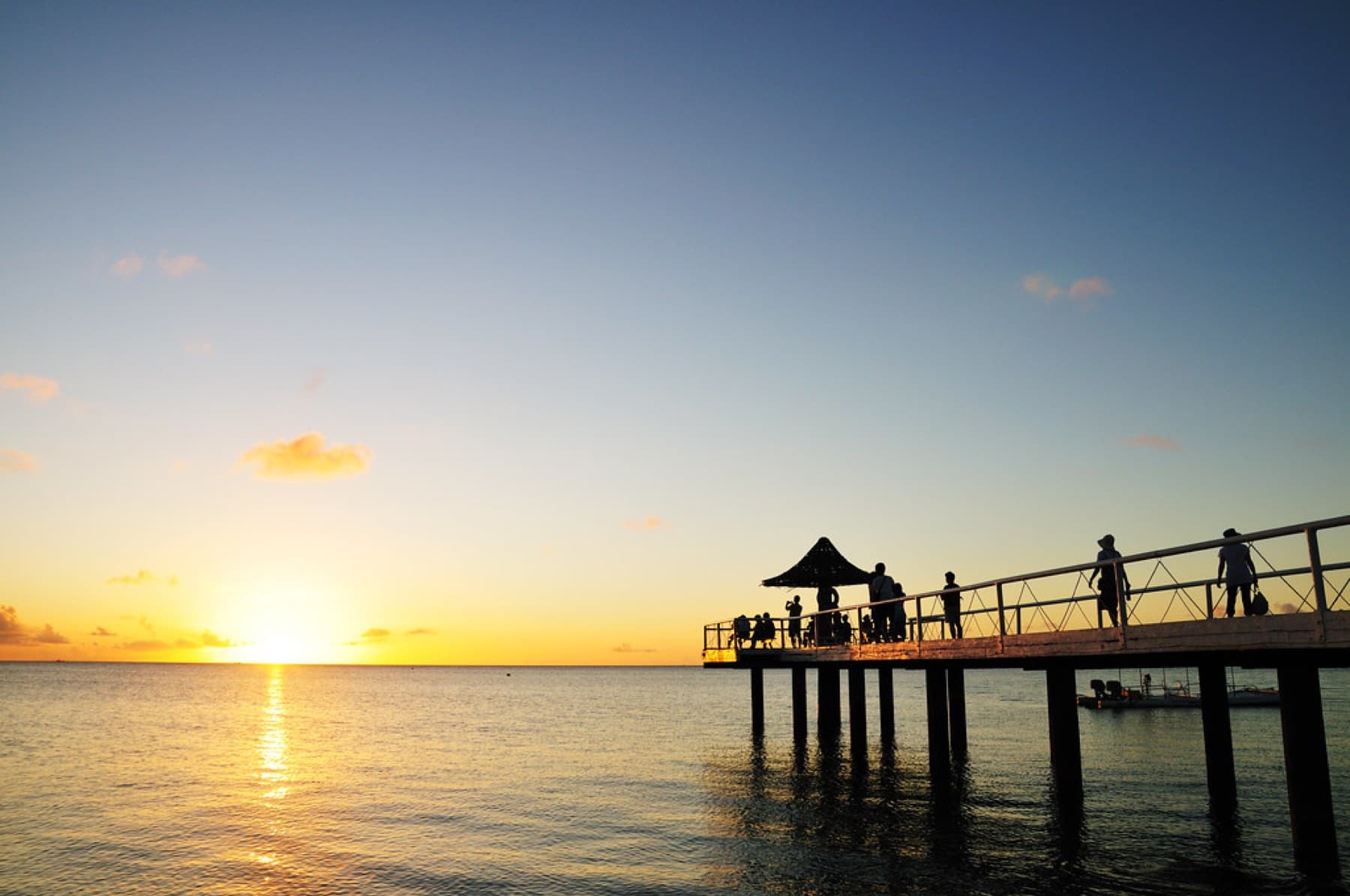 The sun drops toward a calm sea beside the long pier at Fusaki Beach, where a few people stand under the gazebo at its far end.