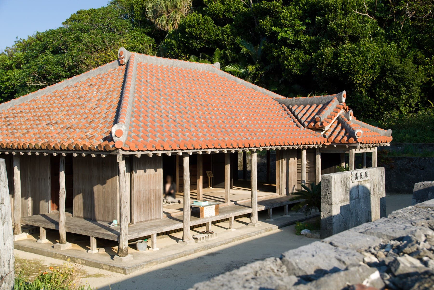 The red-tiled roof of the old Takara house on Geruma rests on wooden posts behind a coral-stone wall, and dense green trees rise beyond it