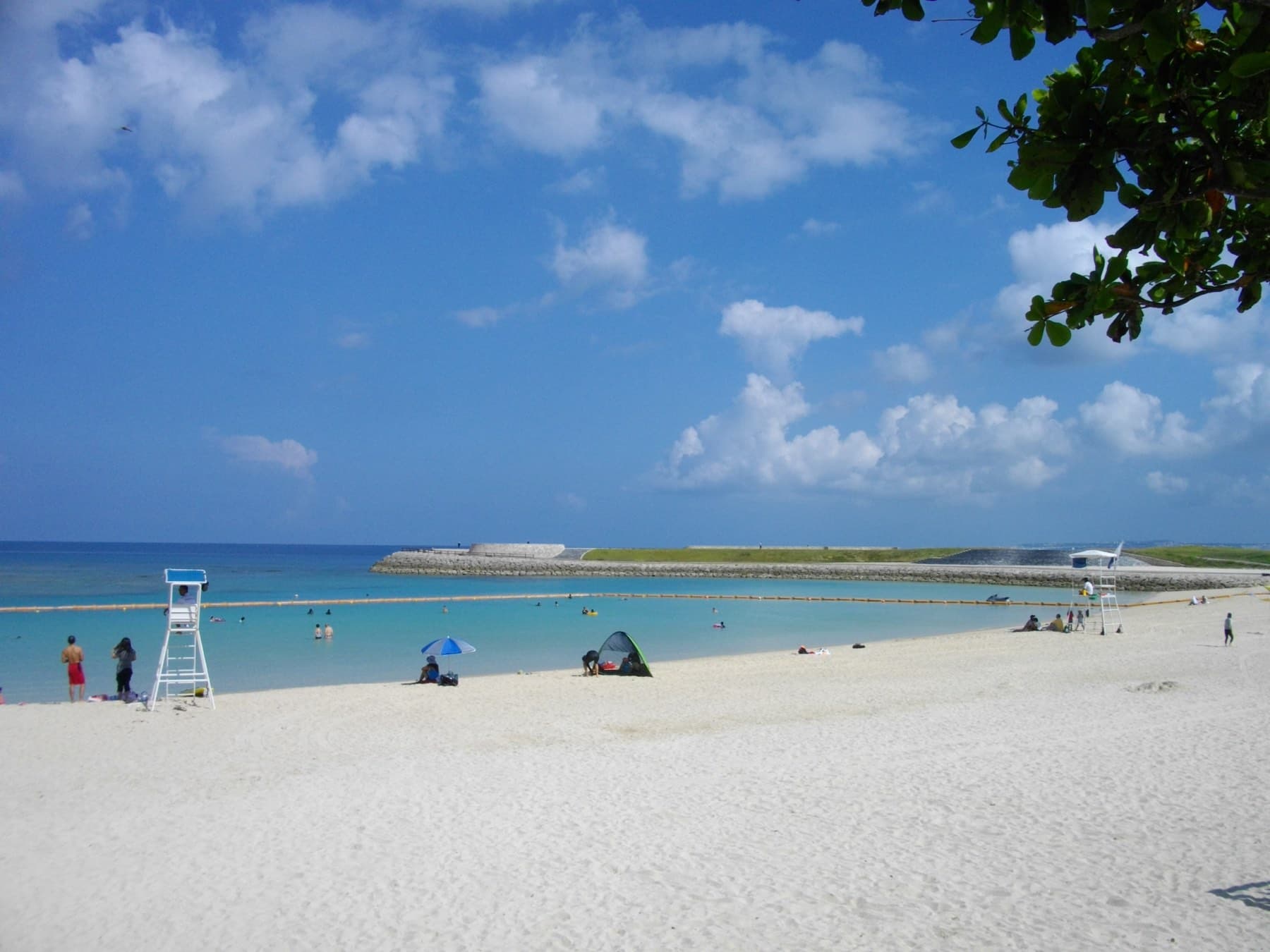 White sand stretches along Ginowan Tropical Beach to a lifeguard tower and a netted swimming area, with a low breakwater across the blue bay