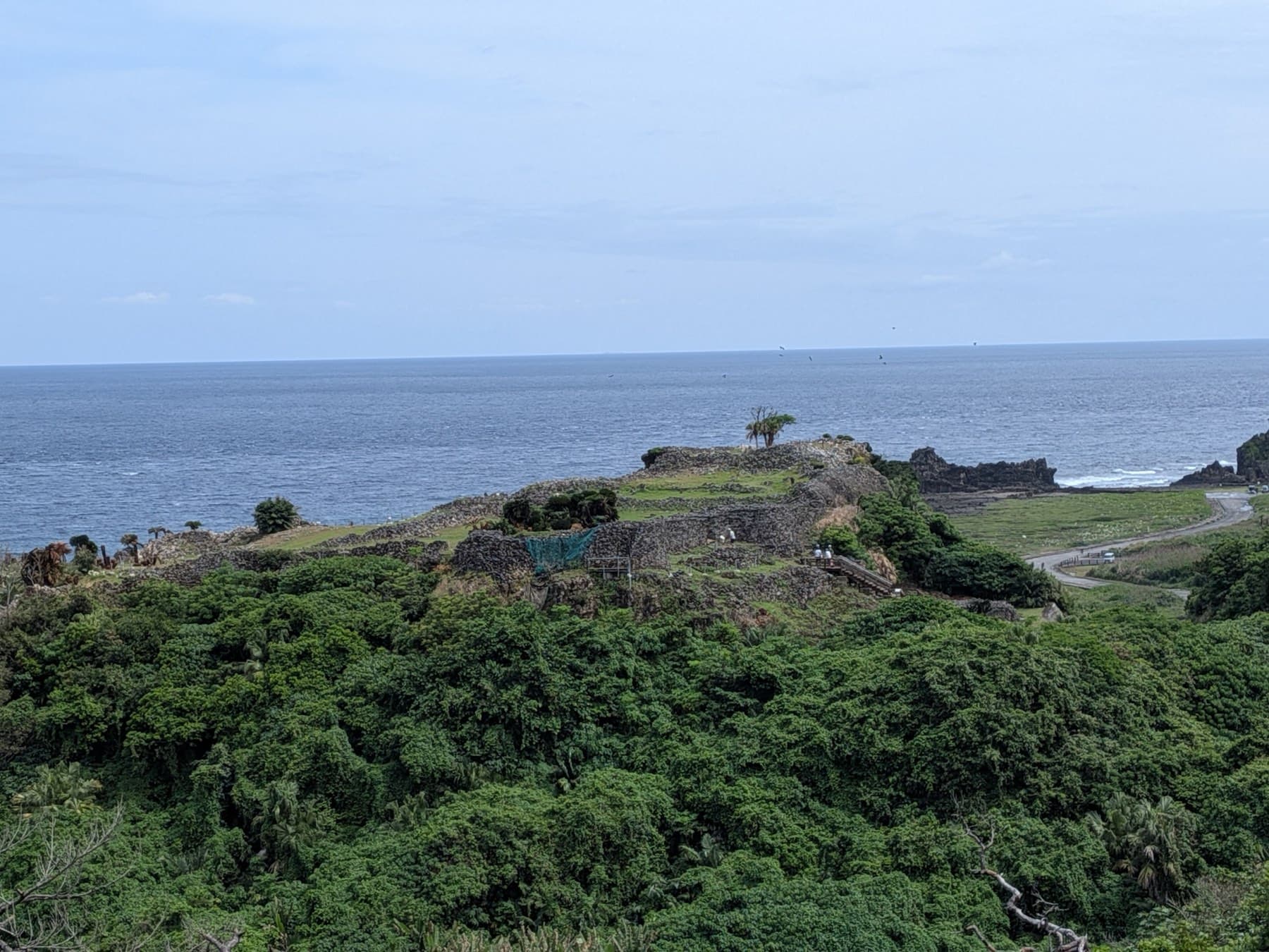 The low stone walls of Gushikawa Castle ruins crown a green clifftop on Kume-jima's north-west coast, with the grey sea stretching to the horizon