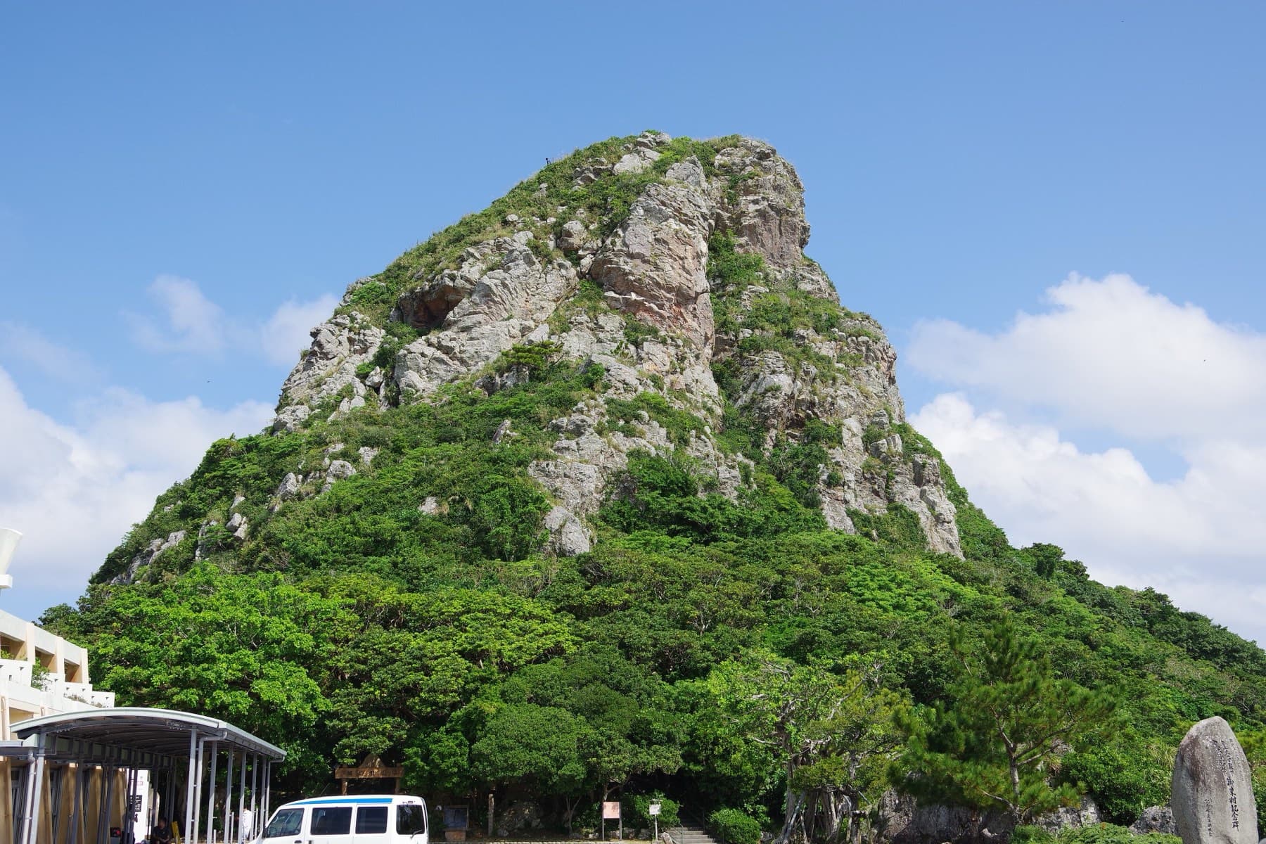 The bare rock summit of Gusuku-yama juts above a skirt of dense green forest on Ie-jima against a bright blue sky with white cloud